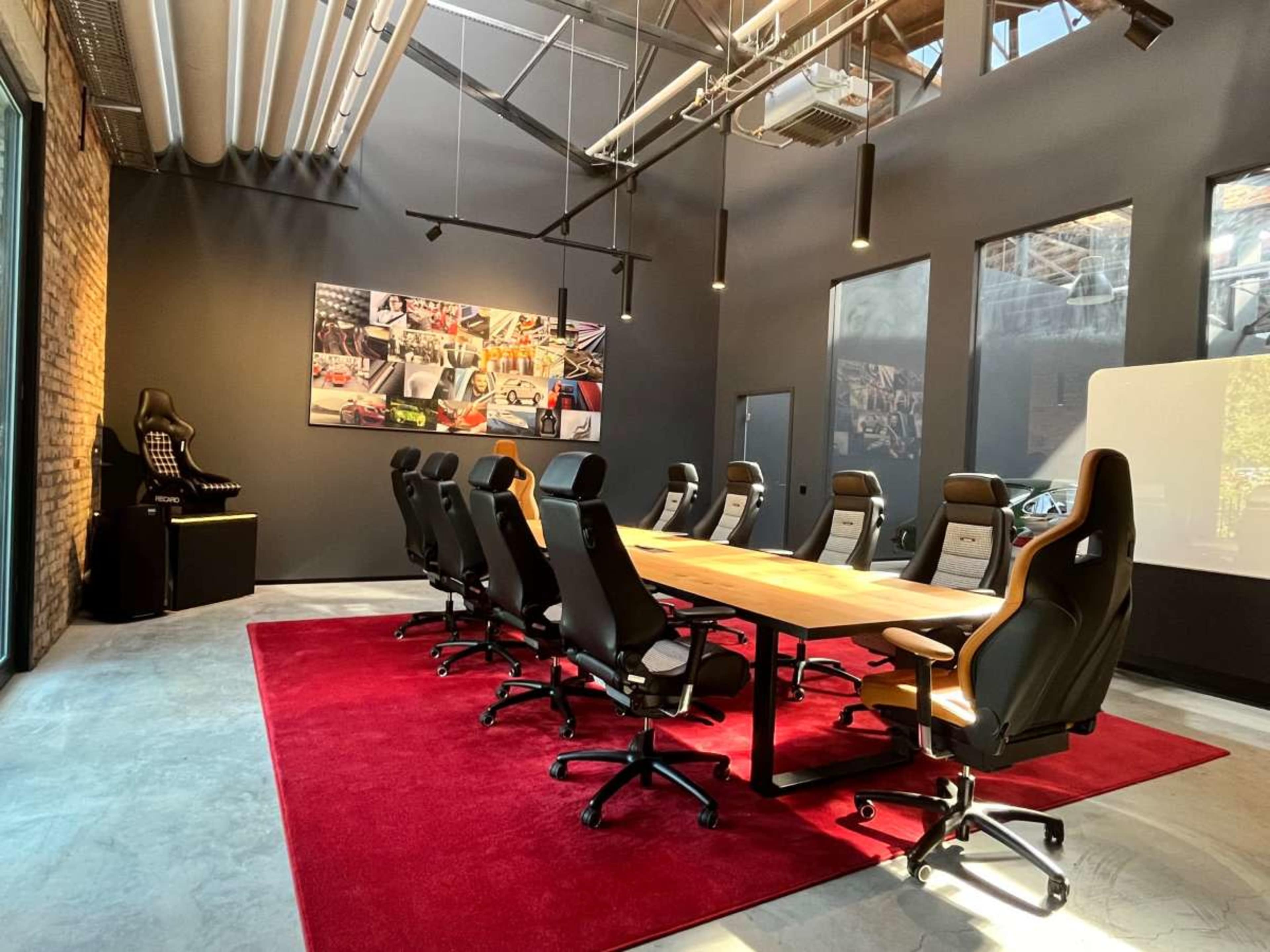 The image depicts a modern conference room with a long wooden table surrounded by eight black office chairs, a red carpet on the floor, and large windows letting in natural light.