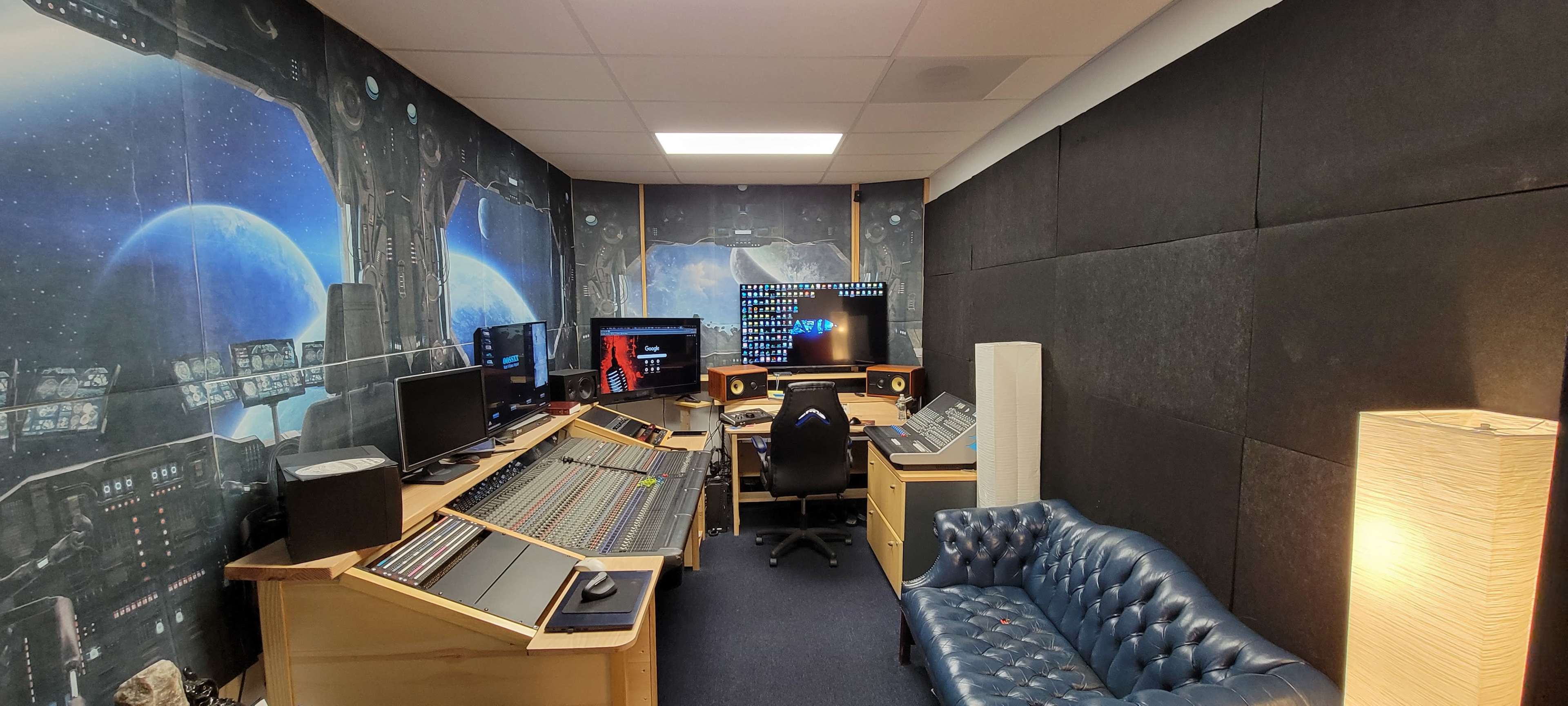 A music production studio featuring multiple computer screens, a mixing console, and a blue sofa against a space-themed mural backdrop.