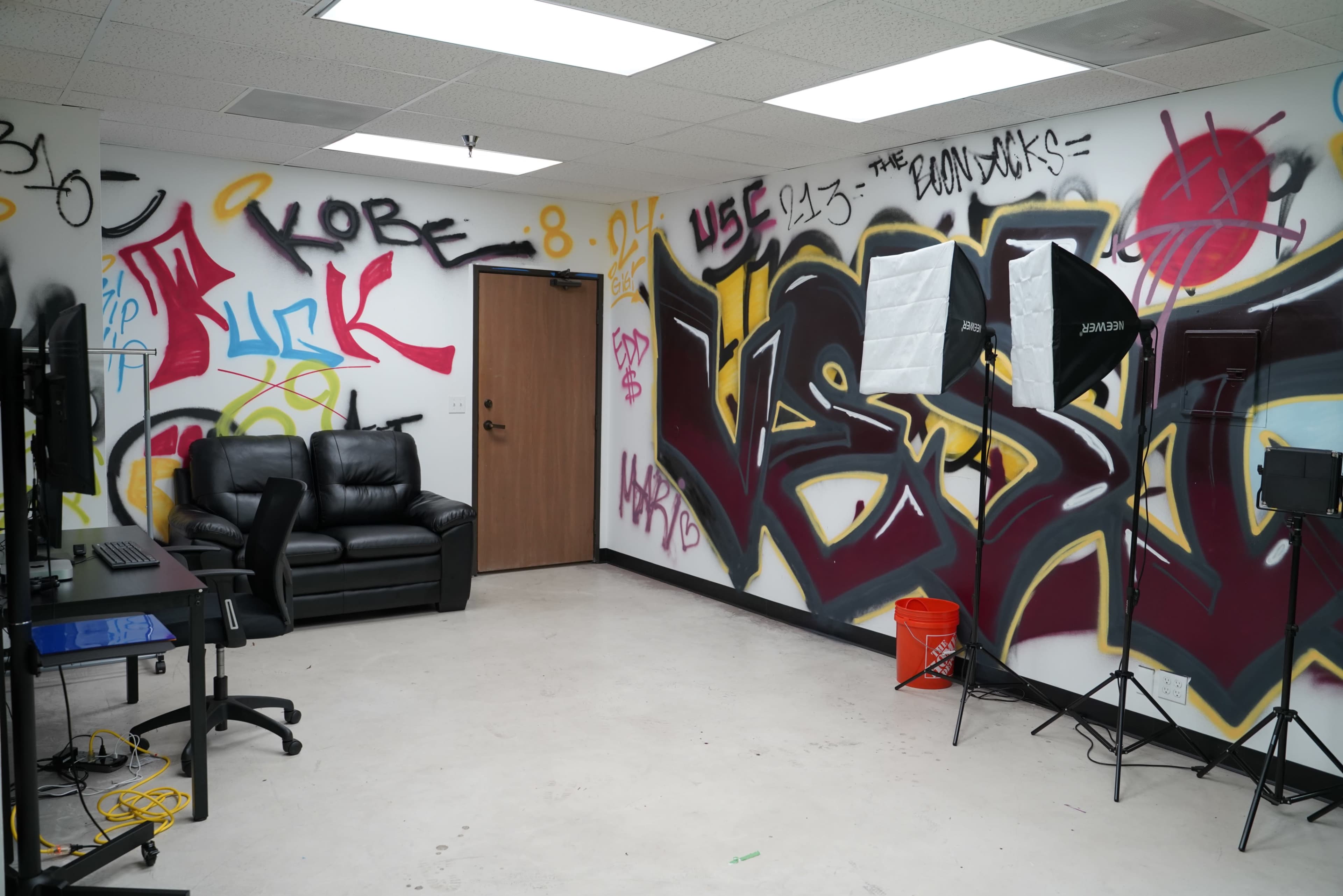 The room features graffiti-covered walls, a black leather couch, and photography lighting equipment set up in a sparse space.