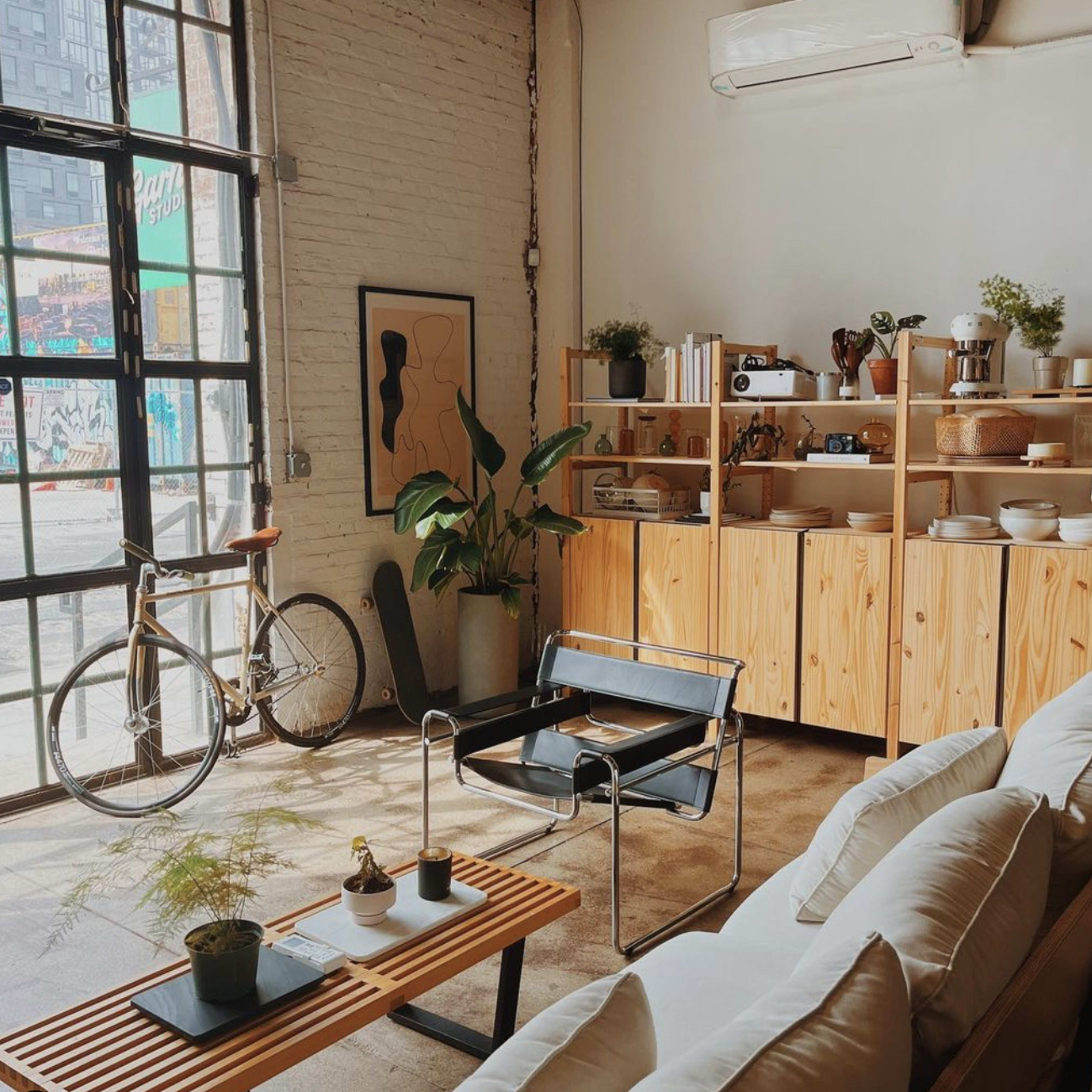 A stylish urban living space featuring a light-colored sofa, a wooden coffee table, and a bike next to a large window with a view of the street outside.