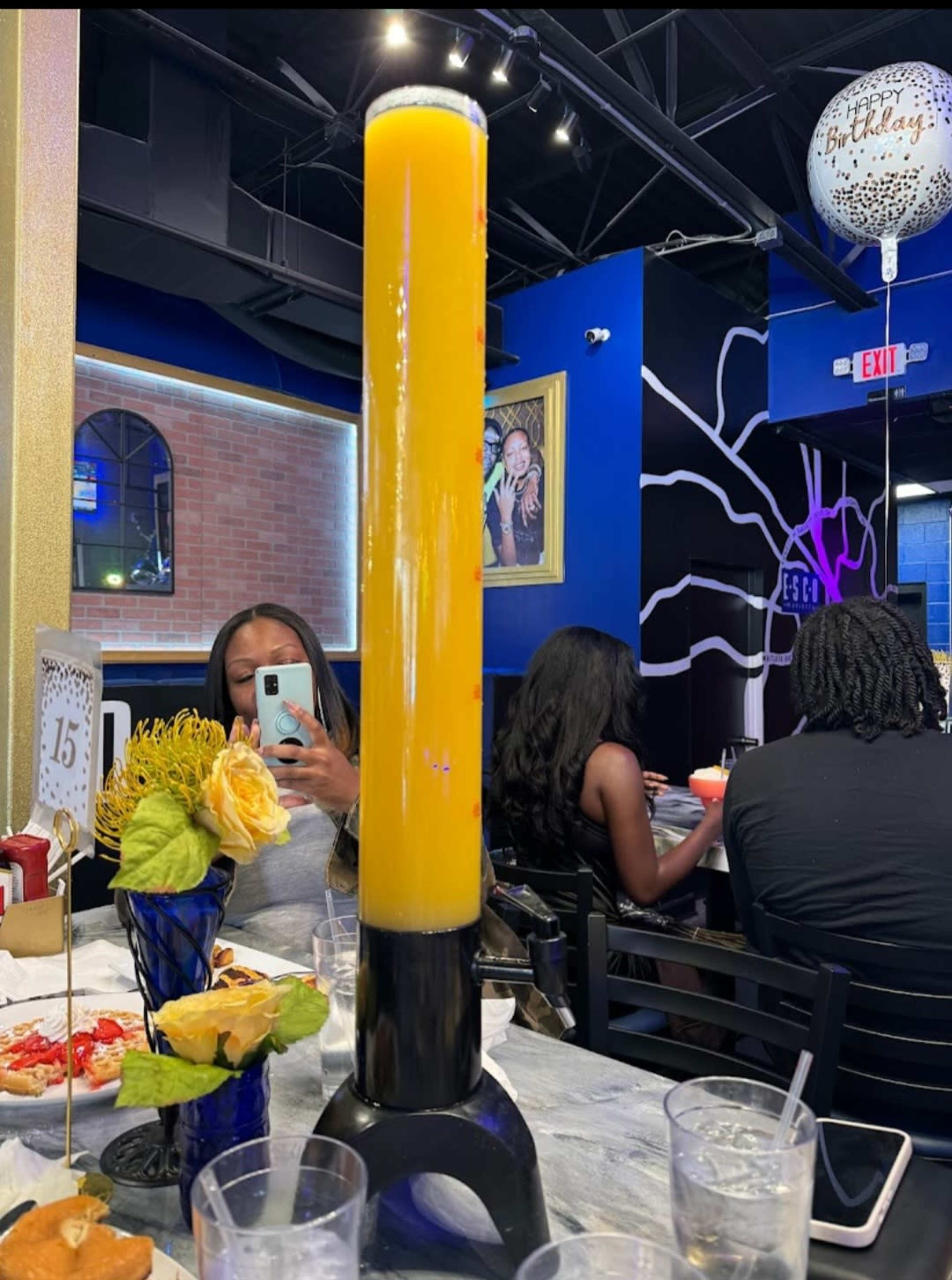 The image shows a restaurant scene featuring a tall drink tower in the foreground, while guests are seated at tables in the background.