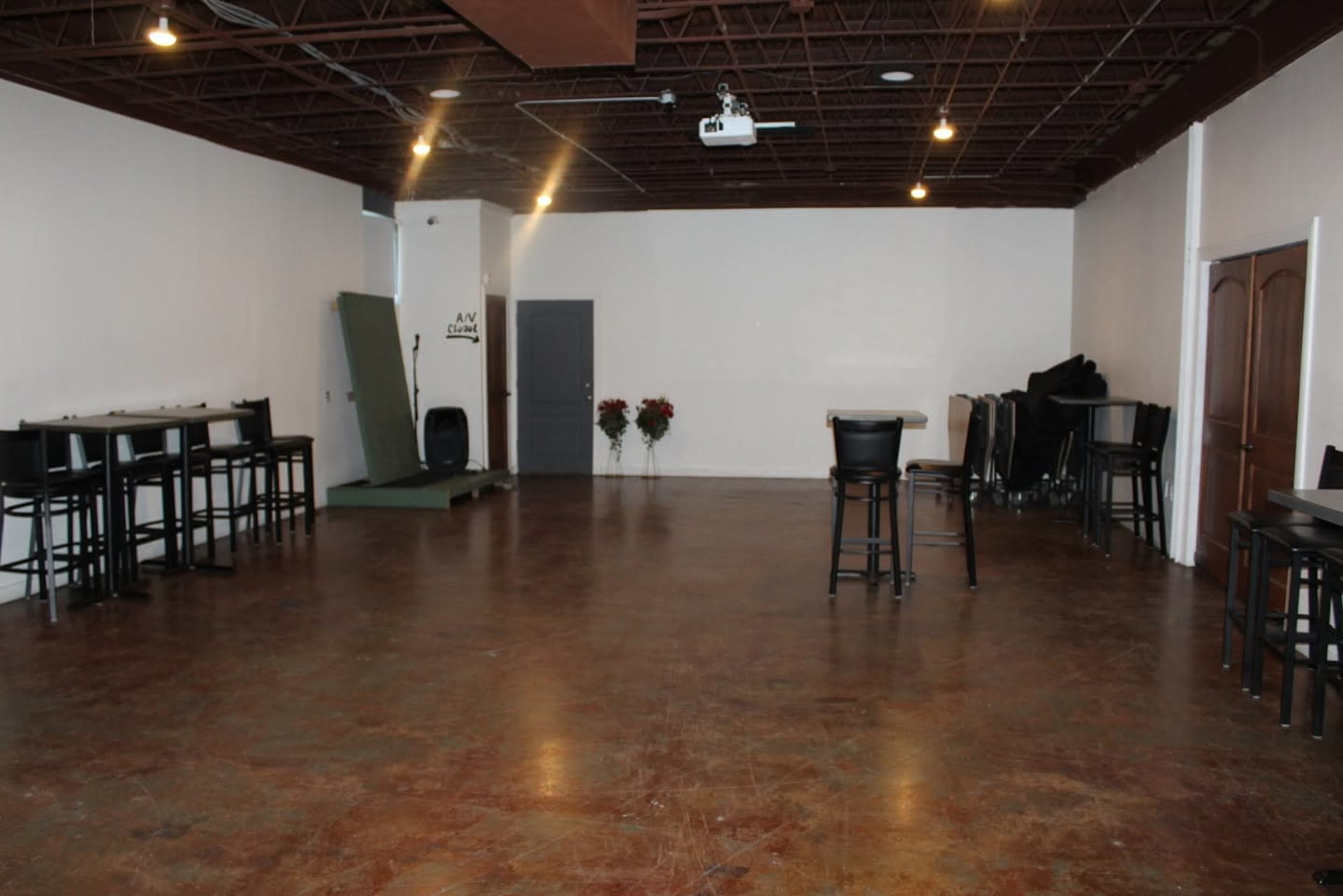 The image shows a spacious, empty room with a polished floor, several tall bar stools arranged around small tables, and a door leading to another area.