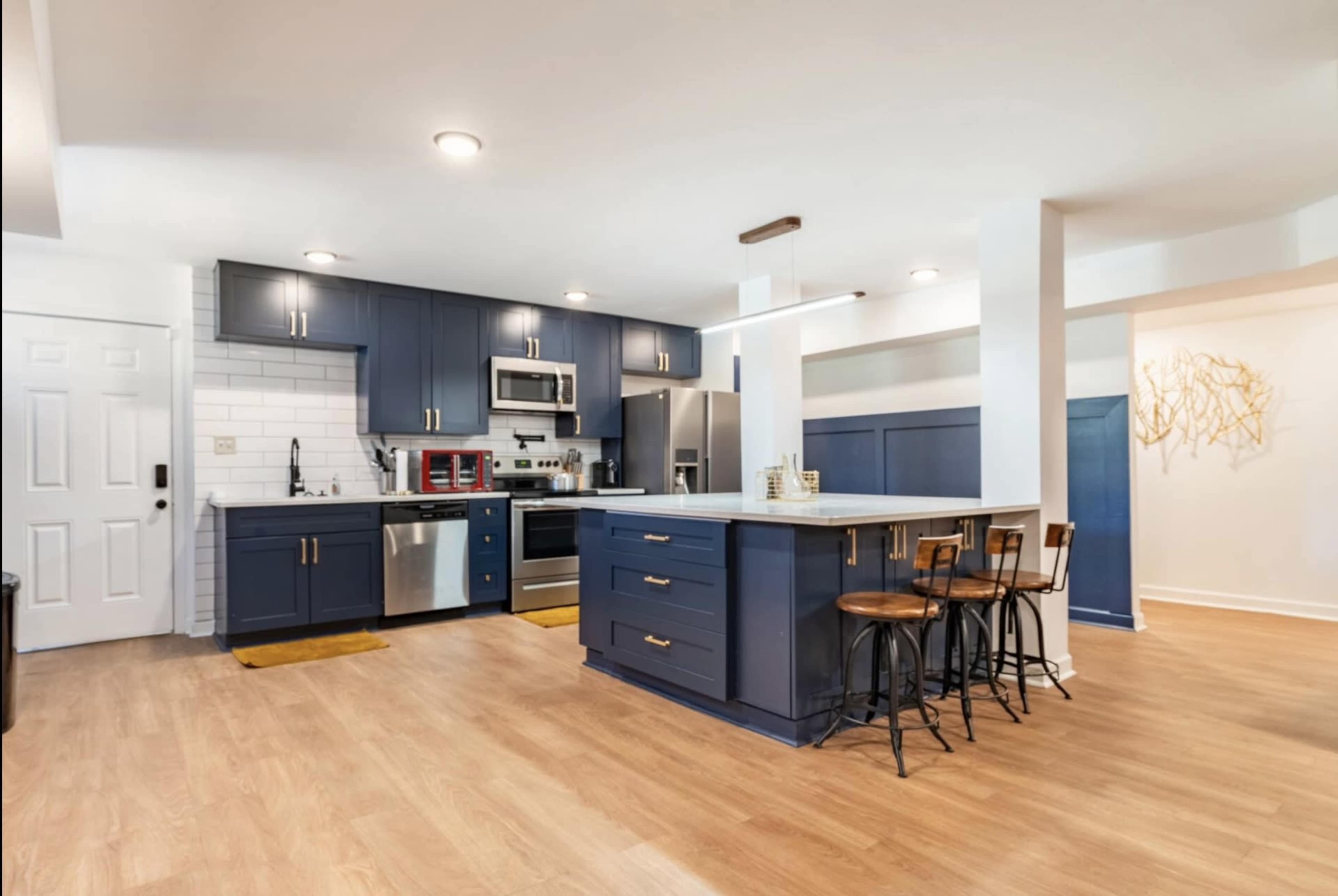 The image shows a modern kitchen featuring navy blue cabinets, stainless steel appliances, and a bar with three stools.