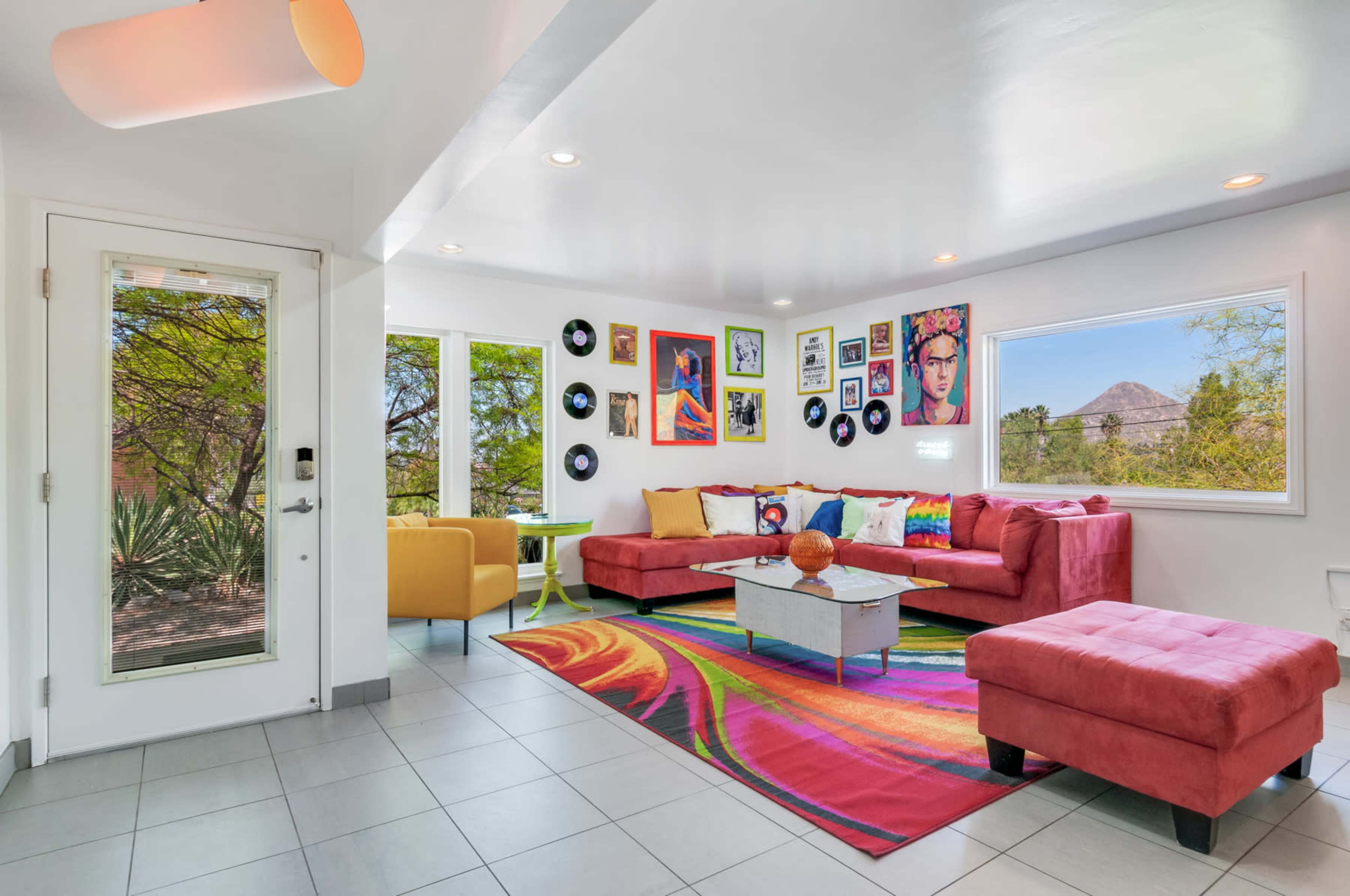 The modern living room features a red sectional sofa, a glass coffee table on a colorful rug, and walls adorned with various framed artworks and vinyl records.