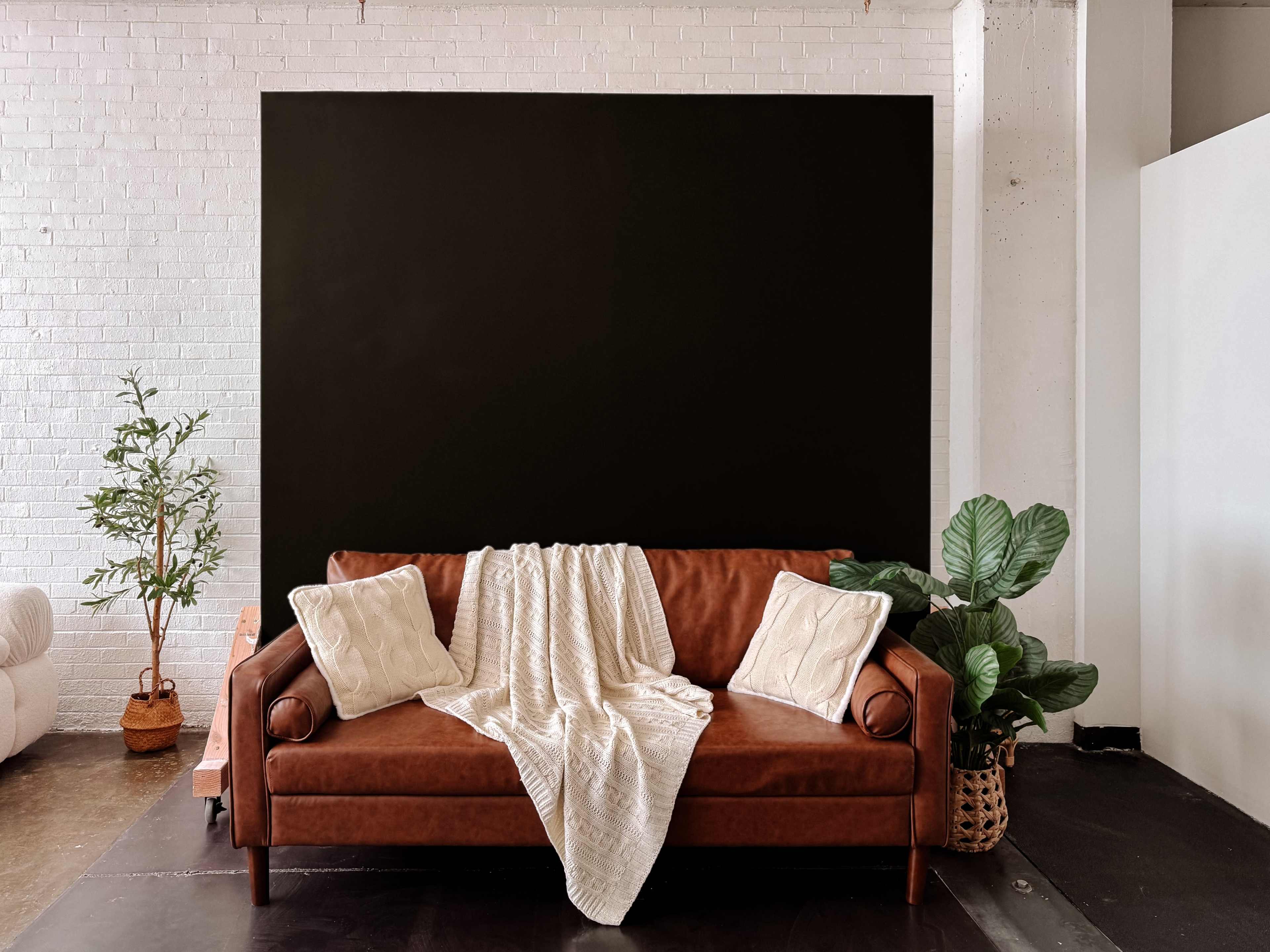 A brown leather sofa with white pillows and a cream-colored throw blanket is placed in front of a large black wall, accompanied by a potted plant on one side.