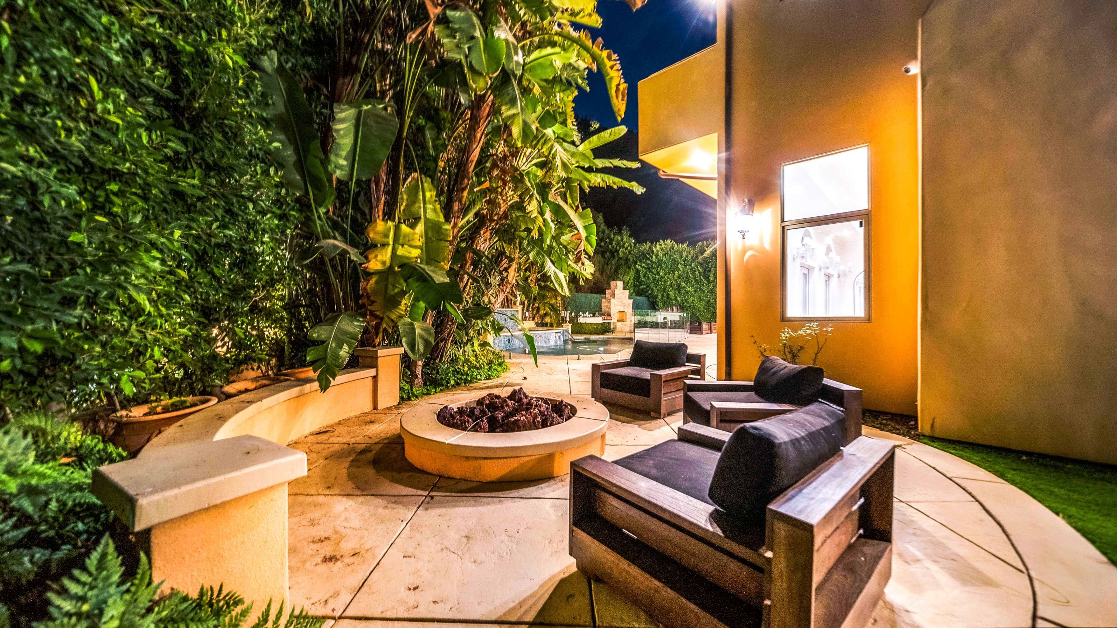 The image displays a cozy outdoor seating area featuring a circular fire pit surrounded by wooden chairs, with lush greenery in the background and a view of a pool.
