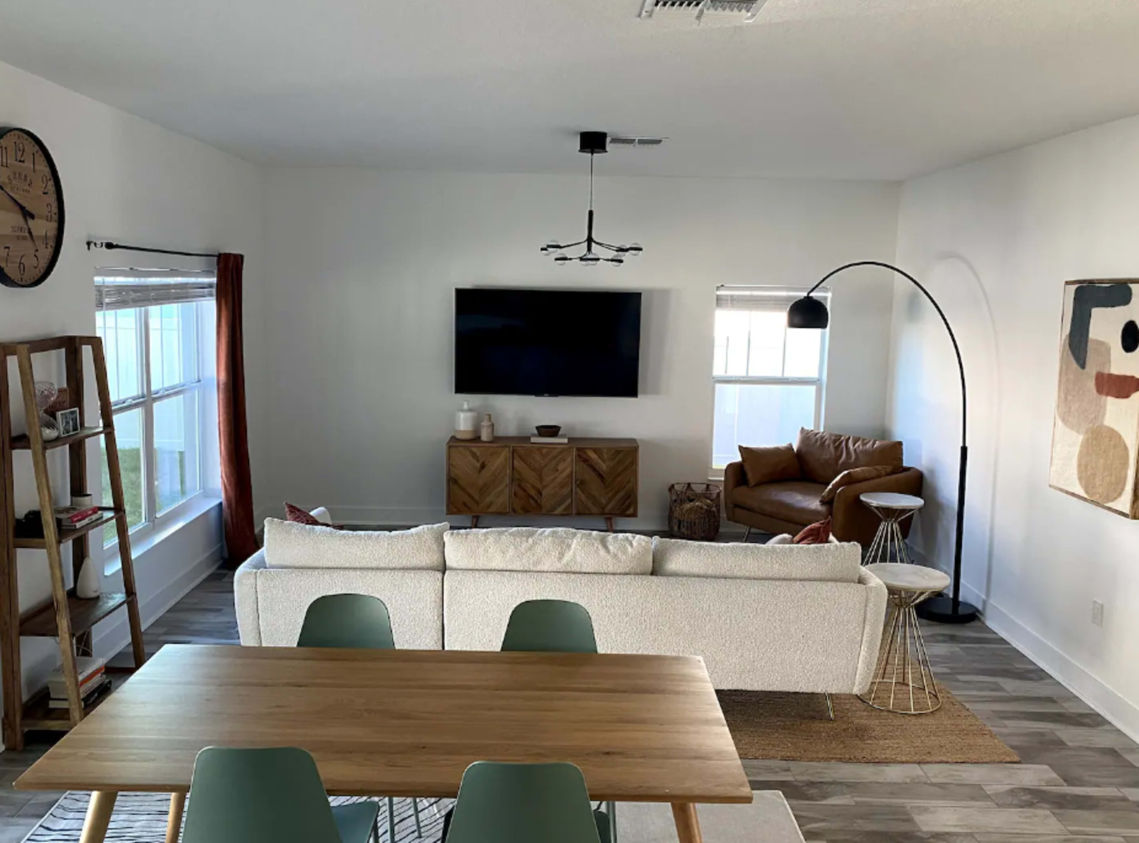 A modern living room features a white sofa, a wooden dining table, a large television on the wall, and a brown leather chair.