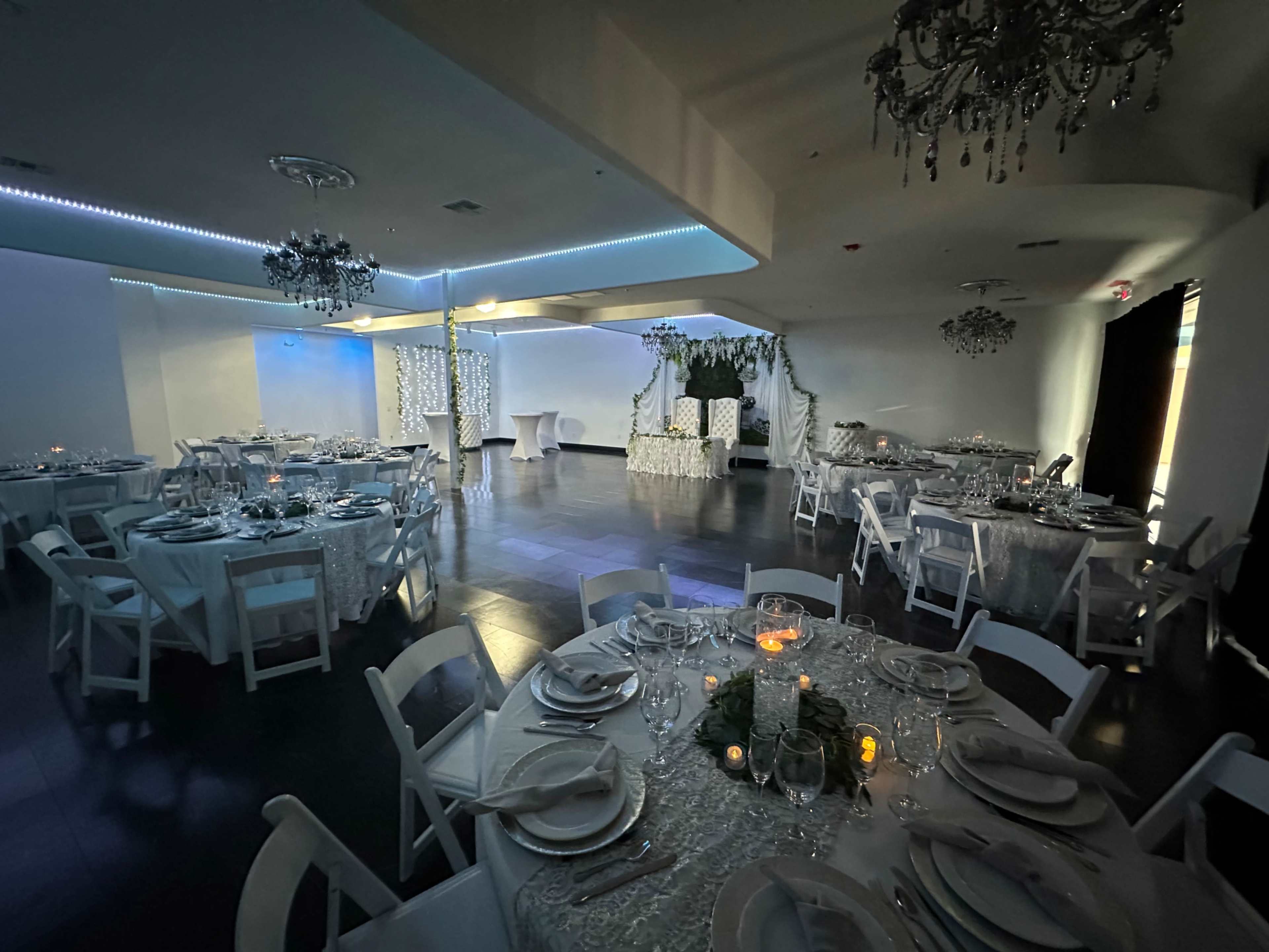 The image shows a decorated event space with circular tables set for dining, illuminated by soft lighting and adorned with floral arrangements.