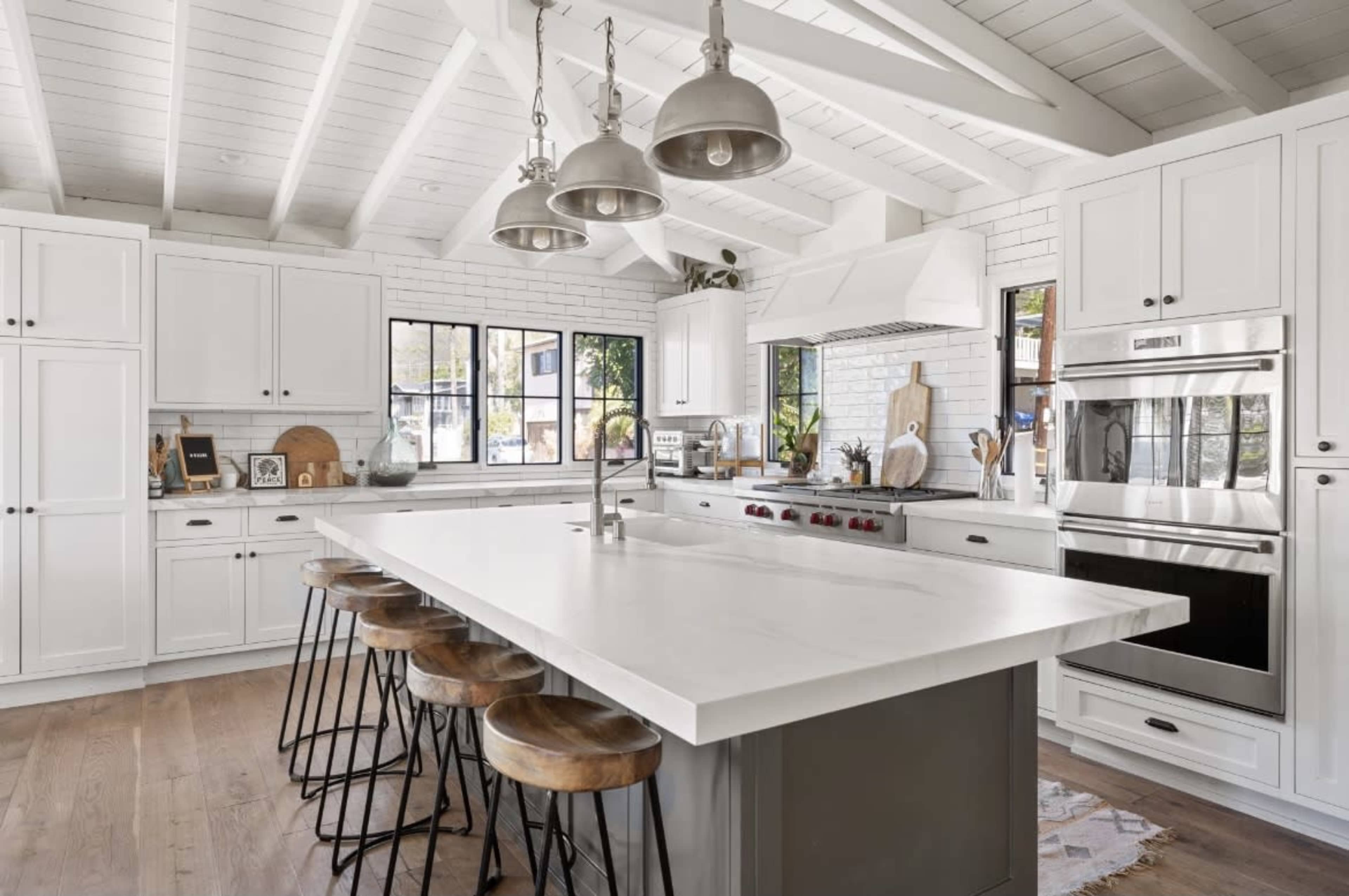 A modern kitchen features a large island with wooden stools, white cabinetry, and stainless steel appliances under a beamed ceiling.
