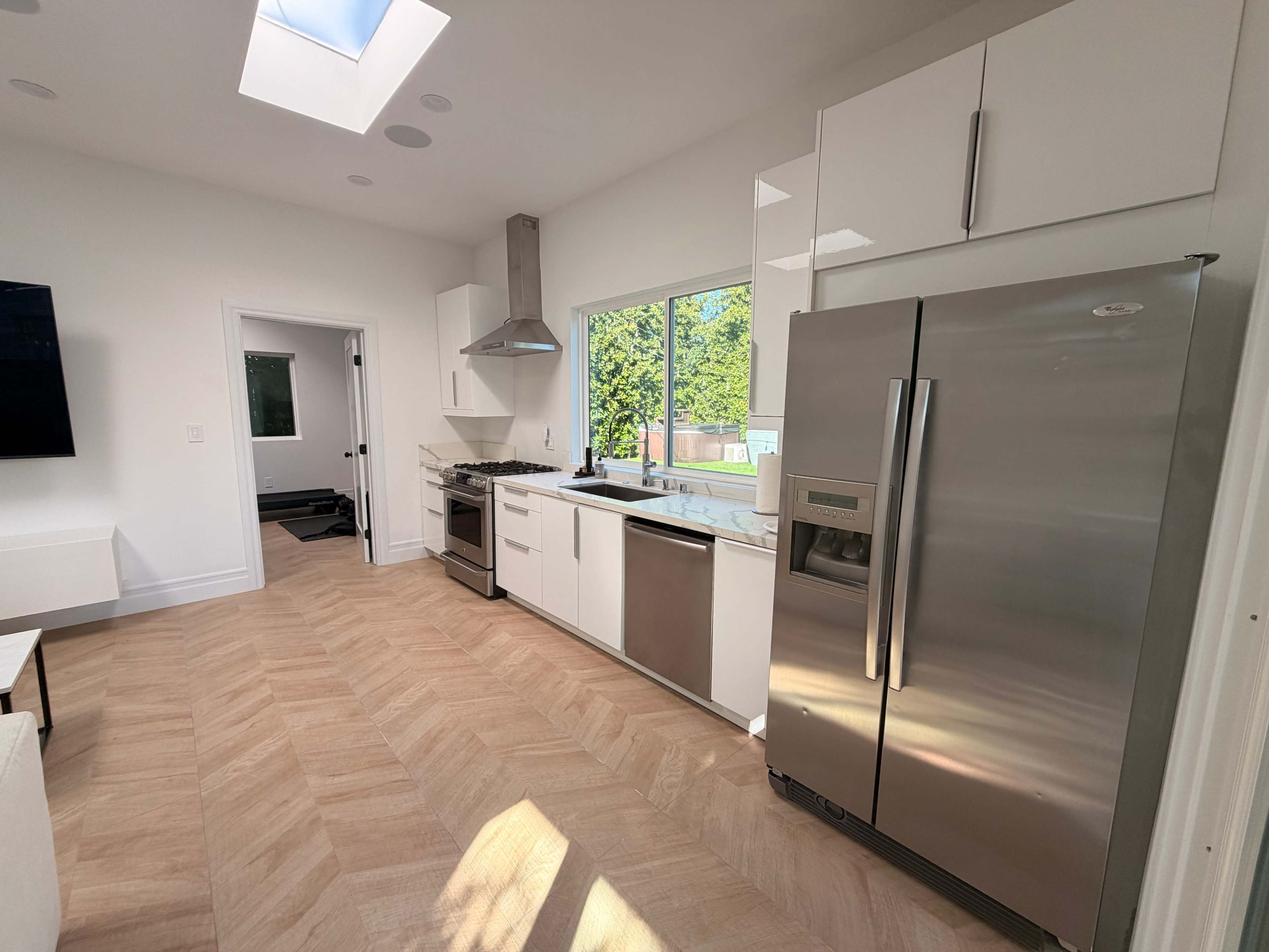 The image shows a modern kitchen with stainless steel appliances, including a refrigerator, oven, and dishwasher, set against white cabinets and a large window.