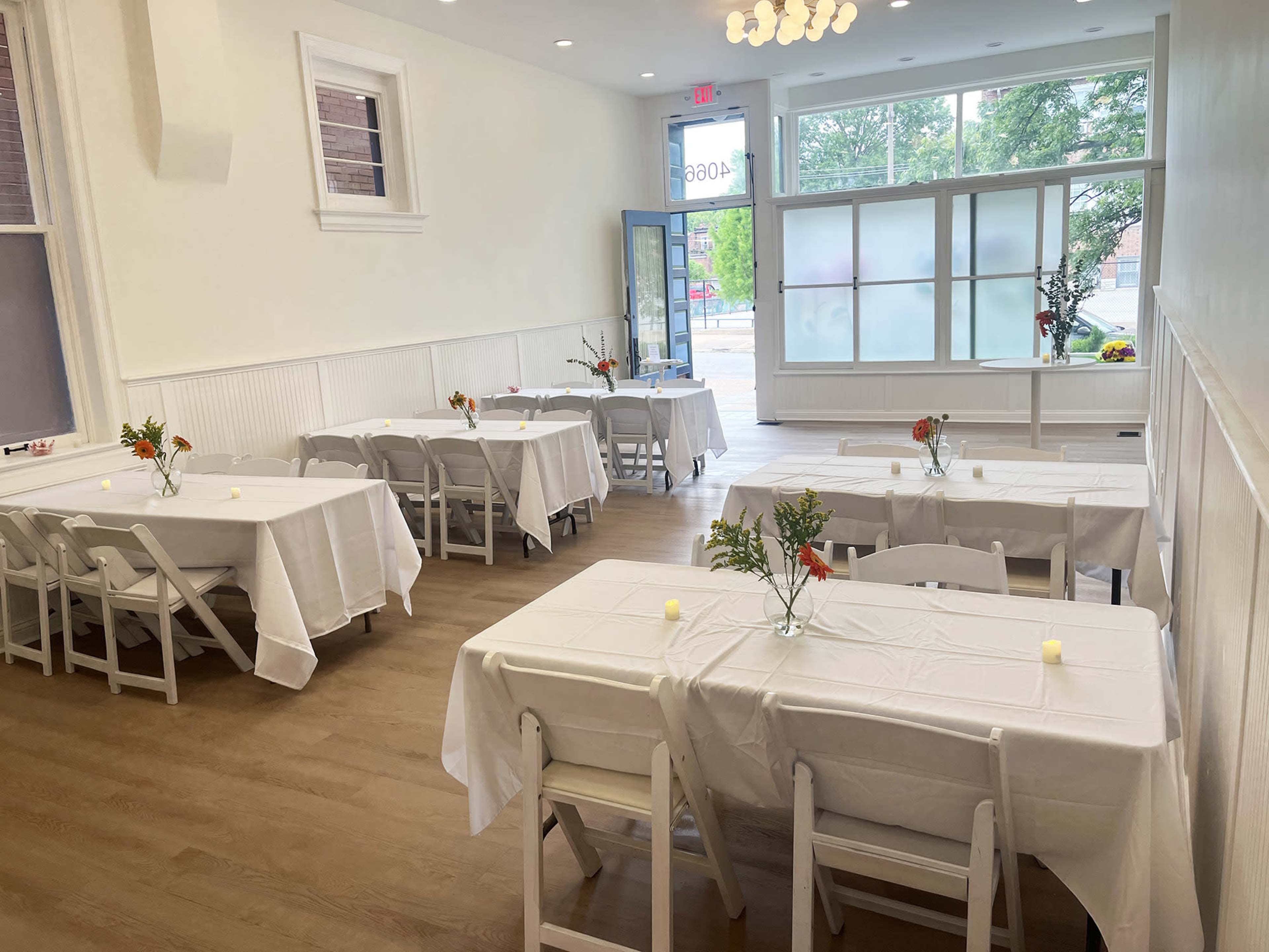 The image shows a bright, organized room with several white tables set for dining, each adorned with a small vase of flowers.