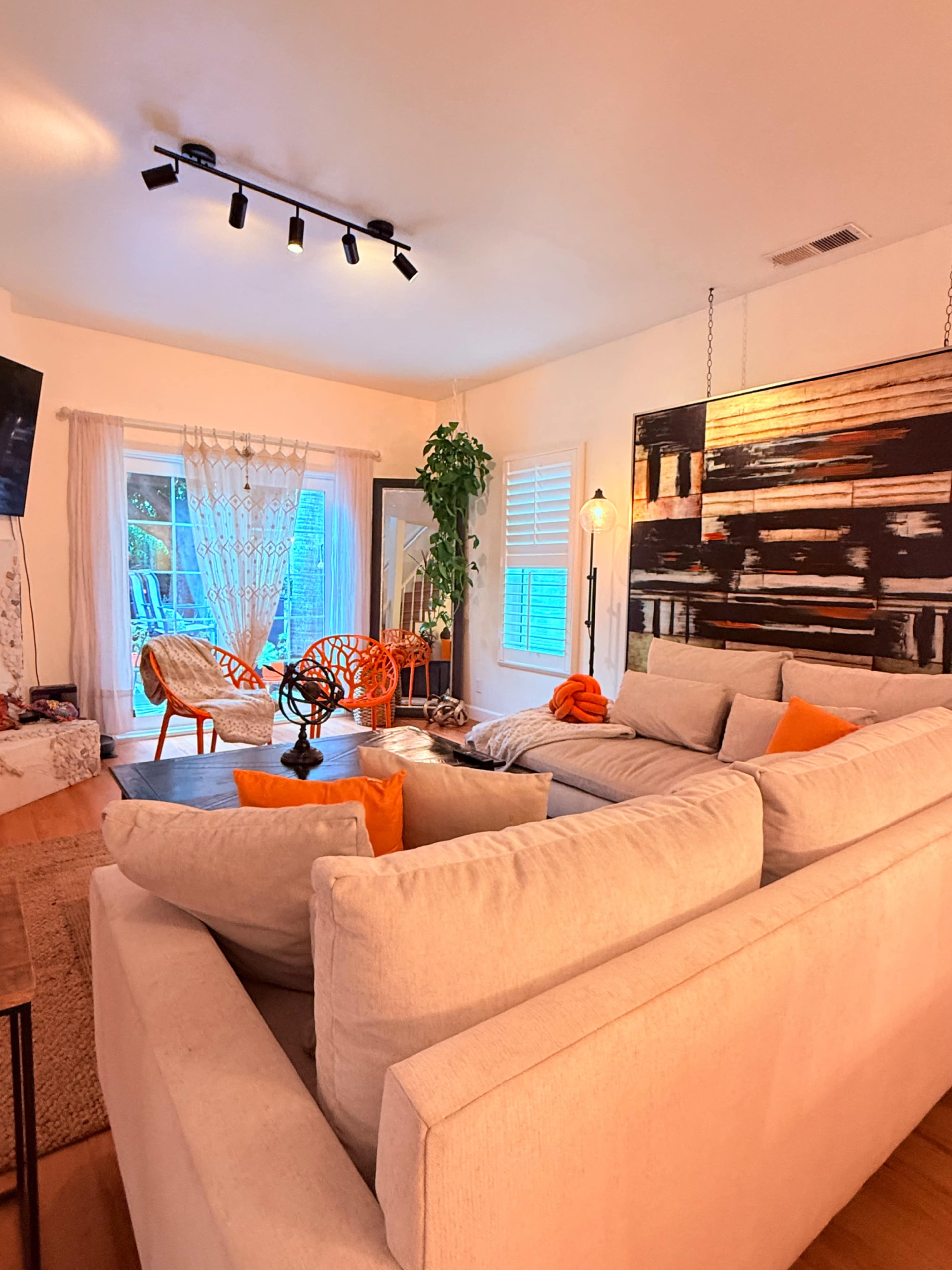 The image shows a modern living room with a large L-shaped sectional sofa, decorative cushions, artwork on the wall, a potted plant, and large windows allowing natural light to stream in.