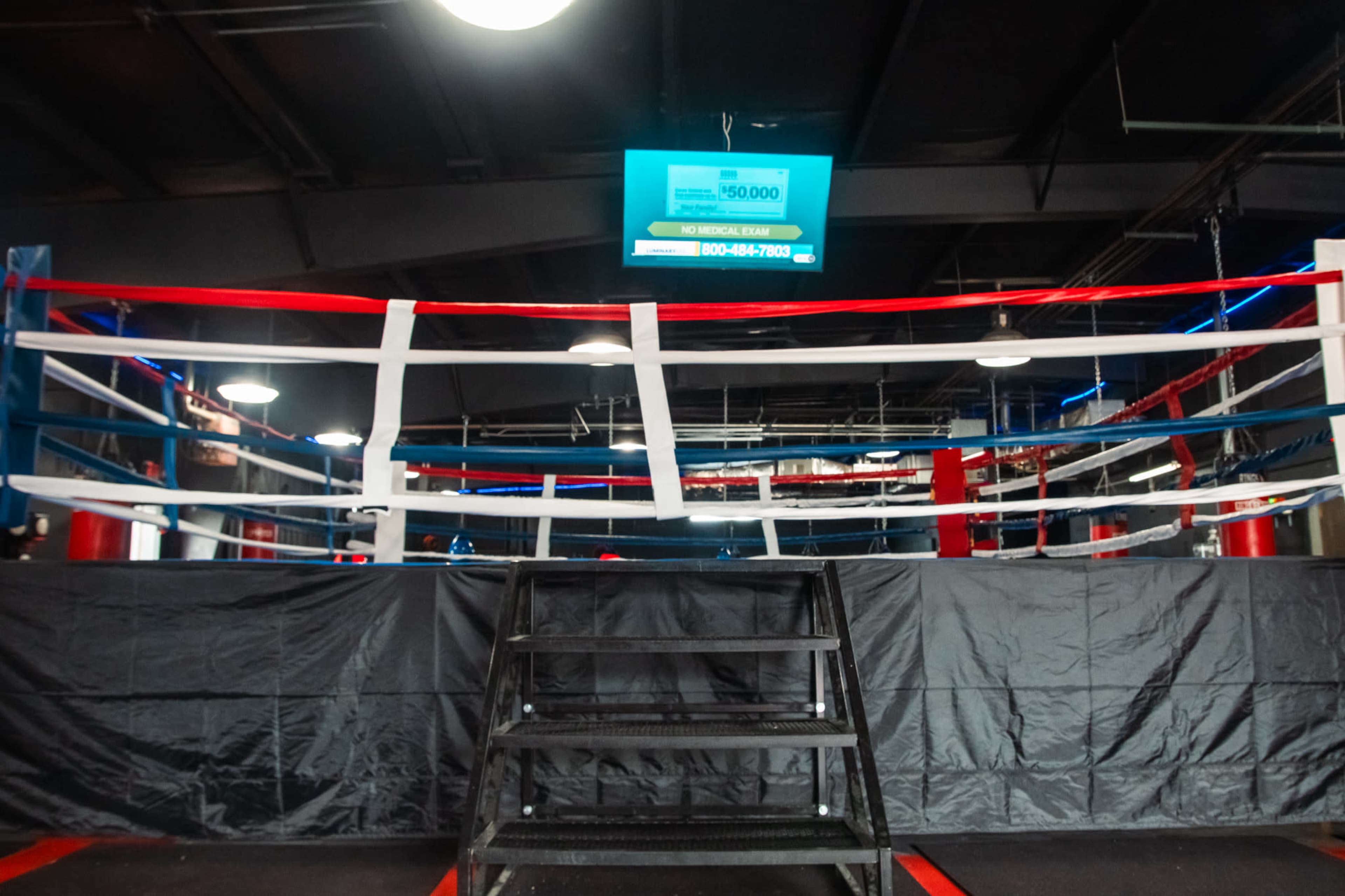 A boxing ring with red, white, and blue ropes is set up in a gym, featuring a staircase leading to the platform.