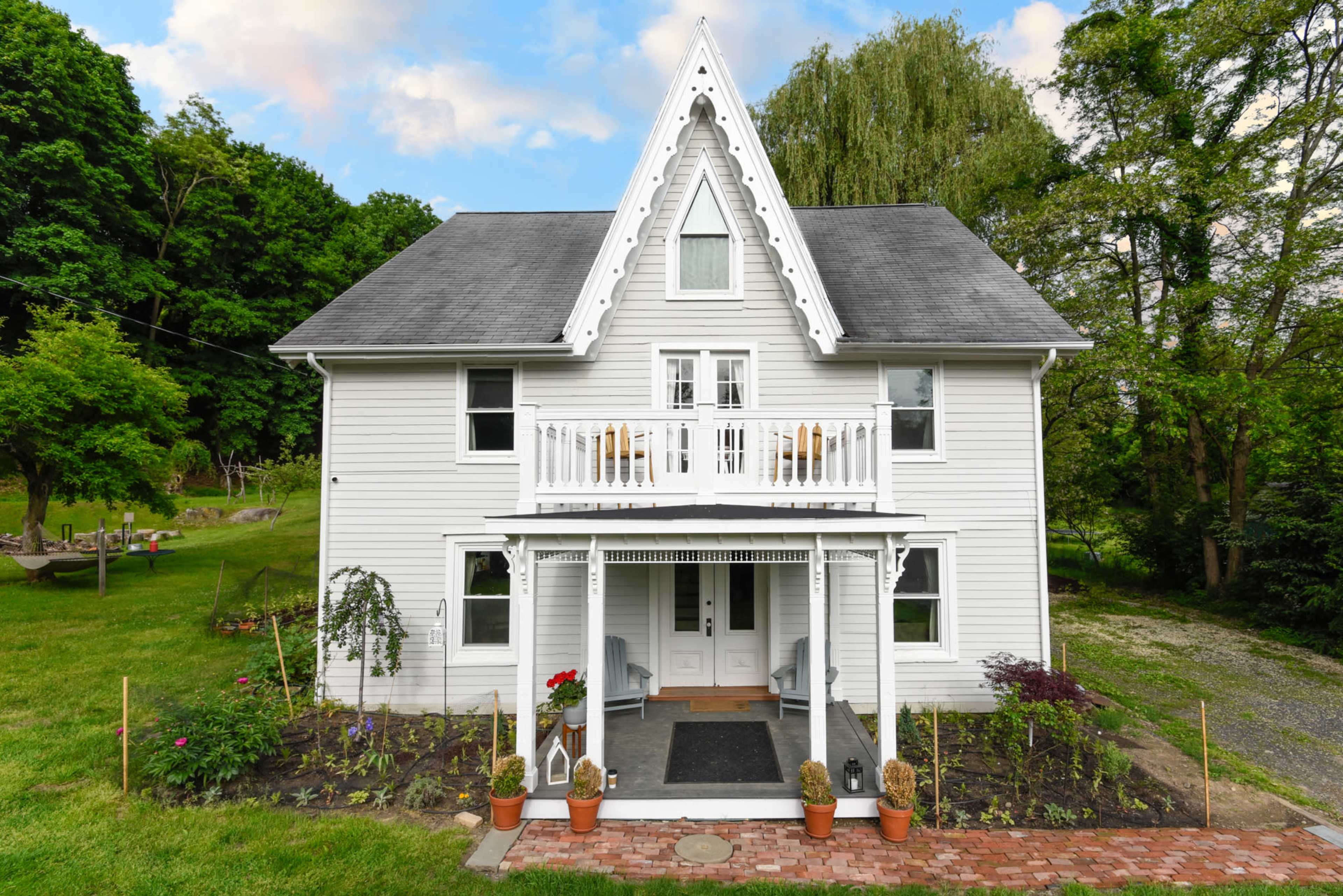 The image shows a white, three-story house with a peaked roof, a front porch, and a grassy yard surrounded by trees.