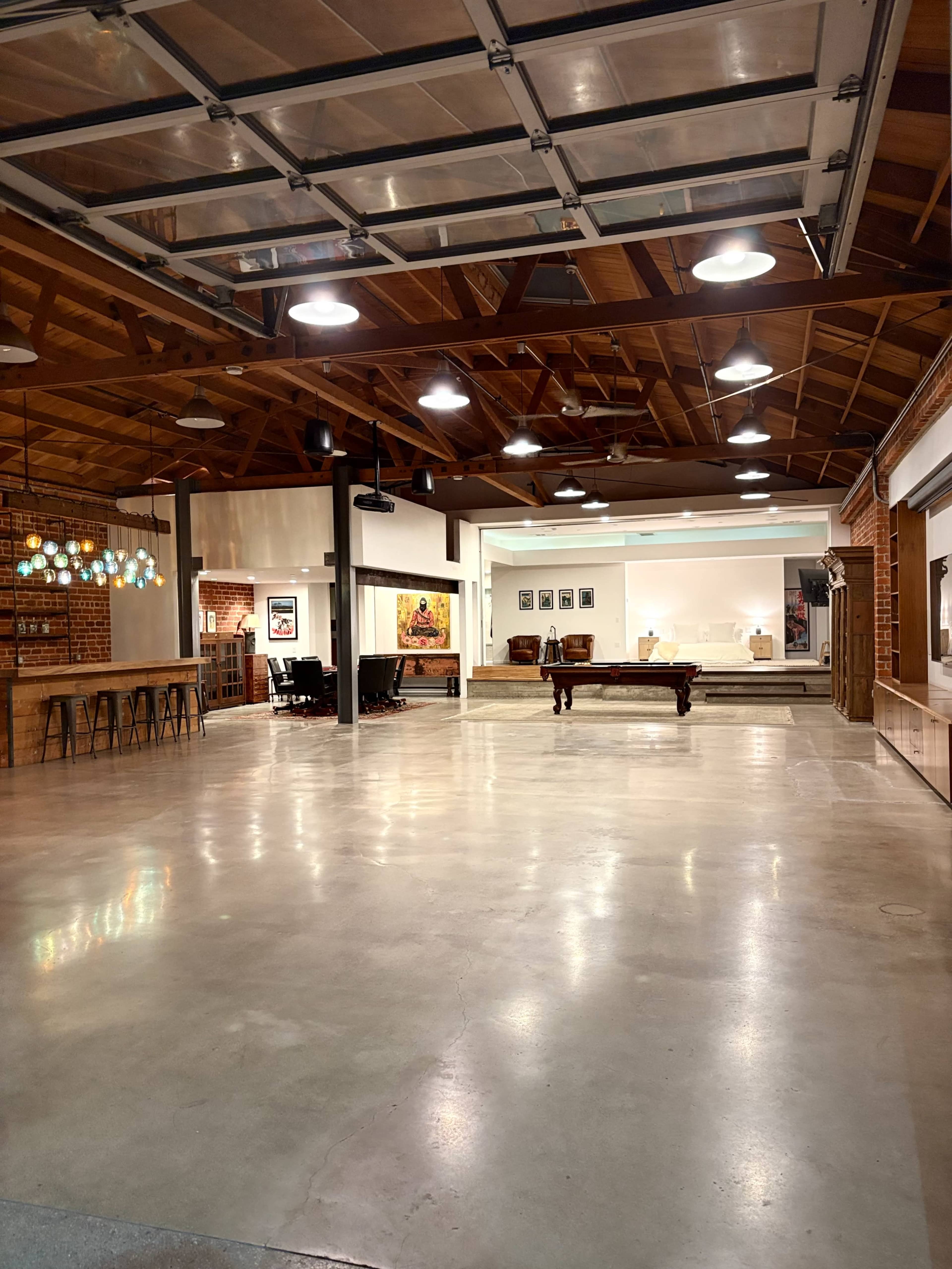 The image shows a spacious and well-lit indoor area with a high wooden ceiling, open floor space, a pool table, and various furniture arrangements.