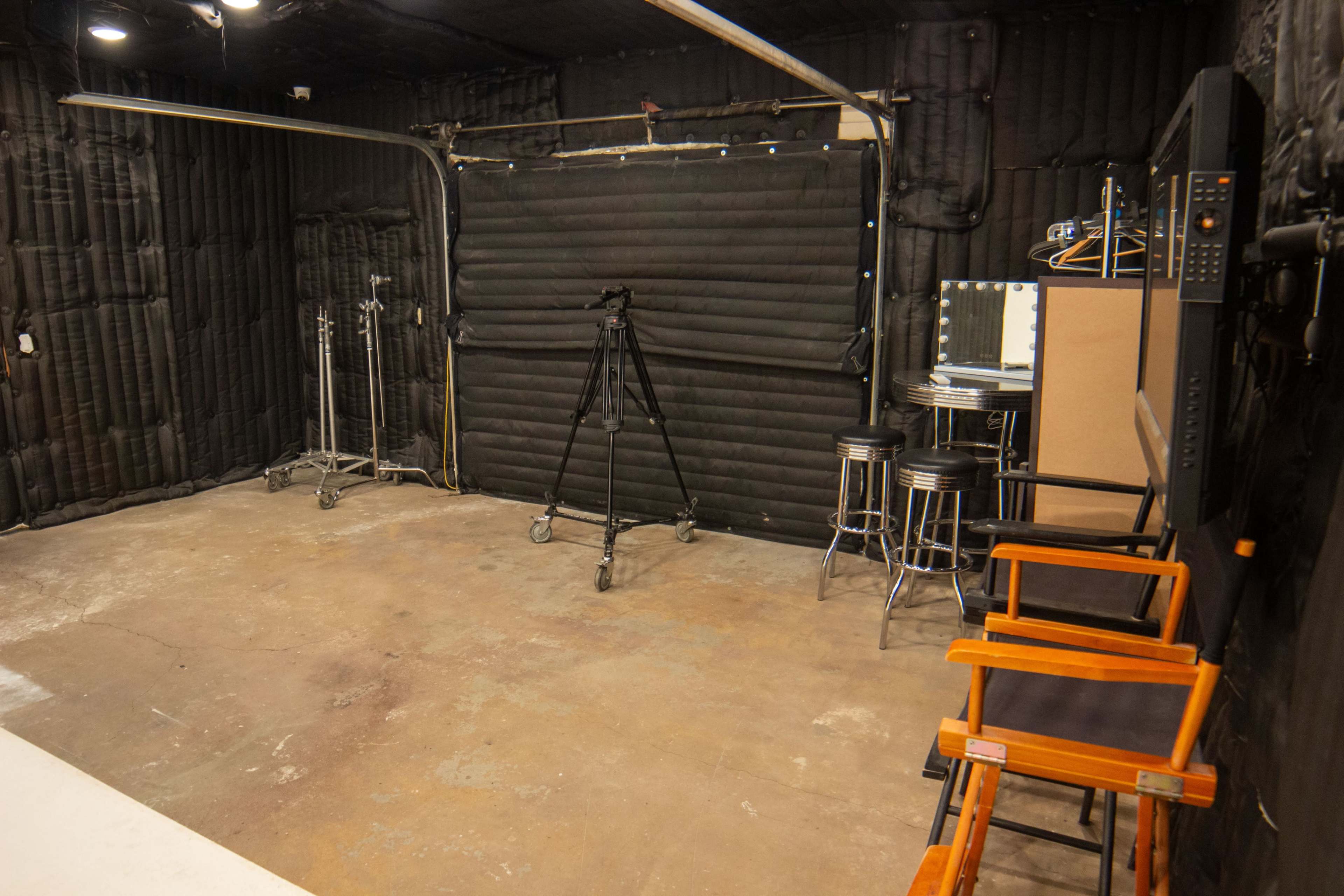 The image shows an empty studio space featuring black padded walls, two tall director's chairs, a mirrored makeup station, and a tripod setup on wheels.