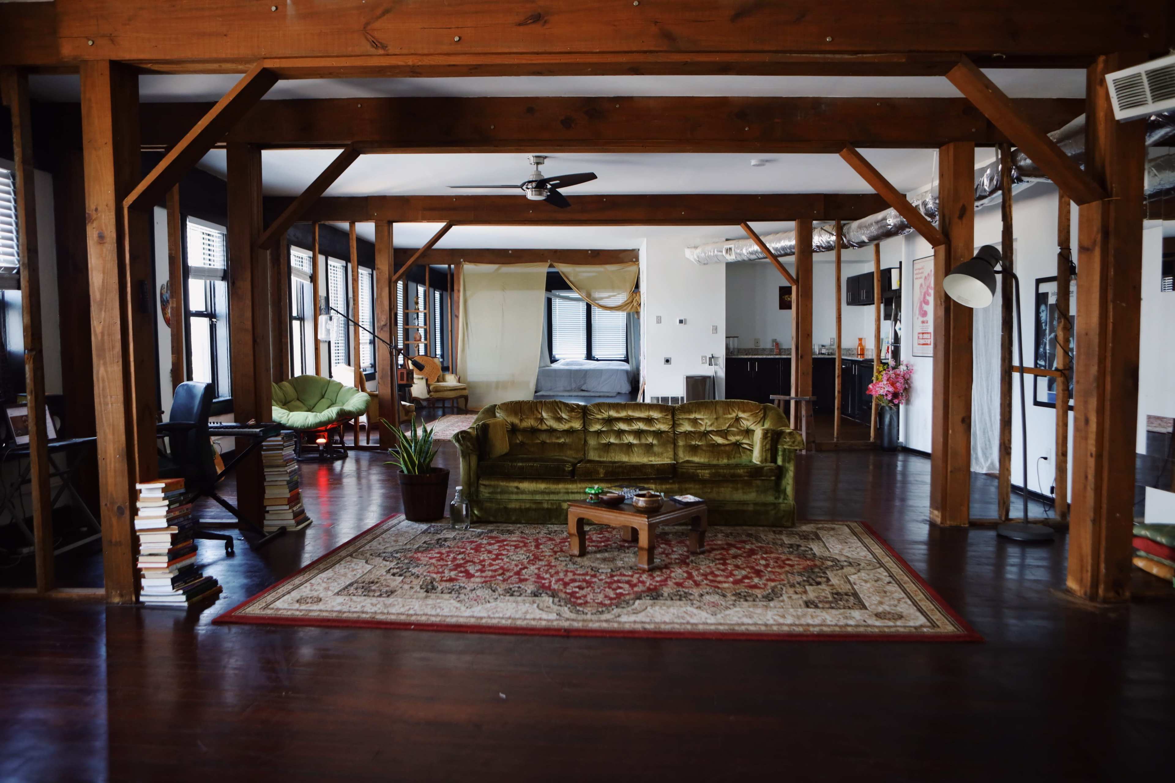A spacious loft-style living area with wooden beams, a green velvet couch, a coffee table, and bookshelves stacked with books.