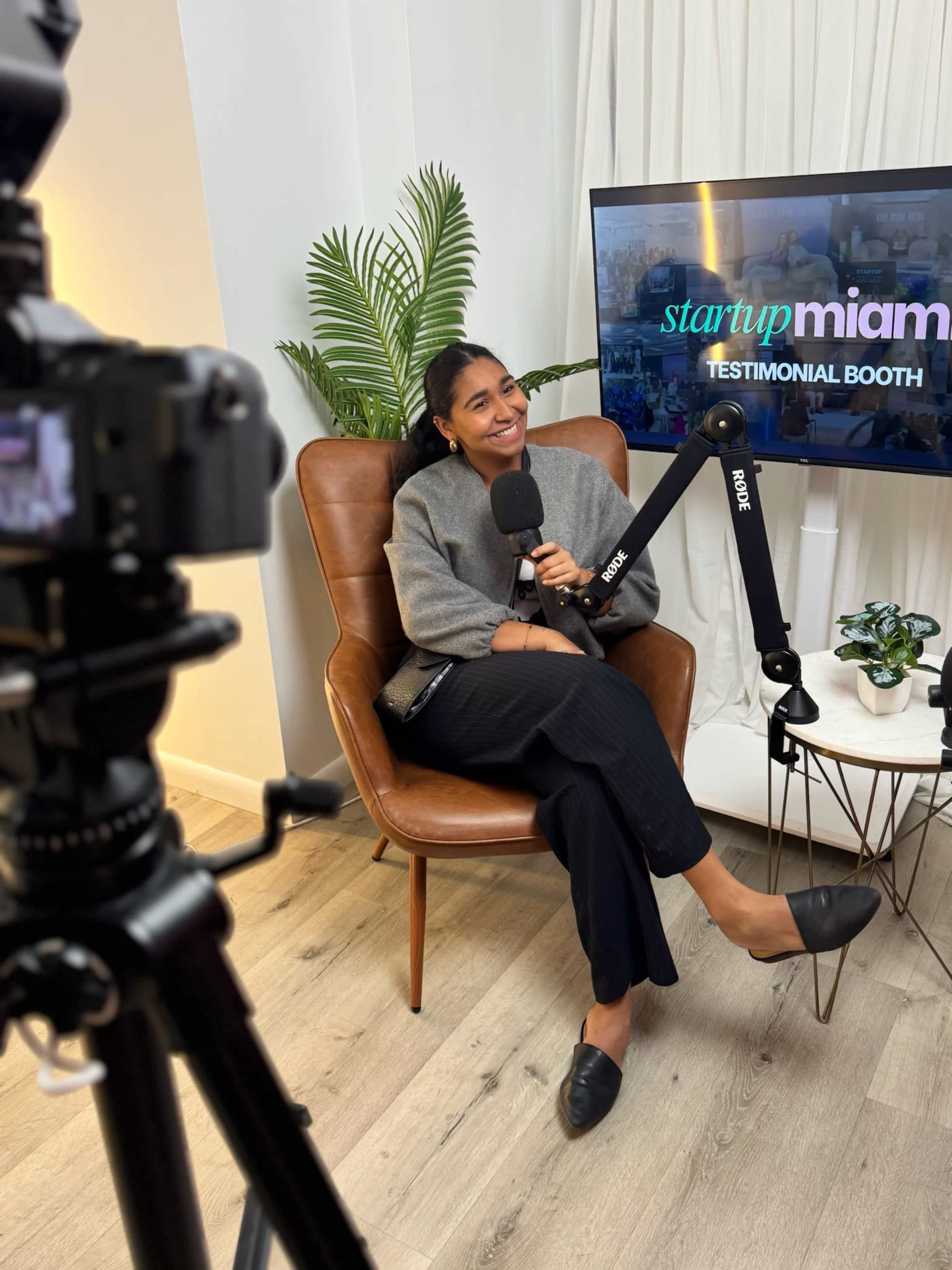 A woman sits in a brown leather chair with a microphone, smiling in front of a screen displaying "startup miami testimonial booth."