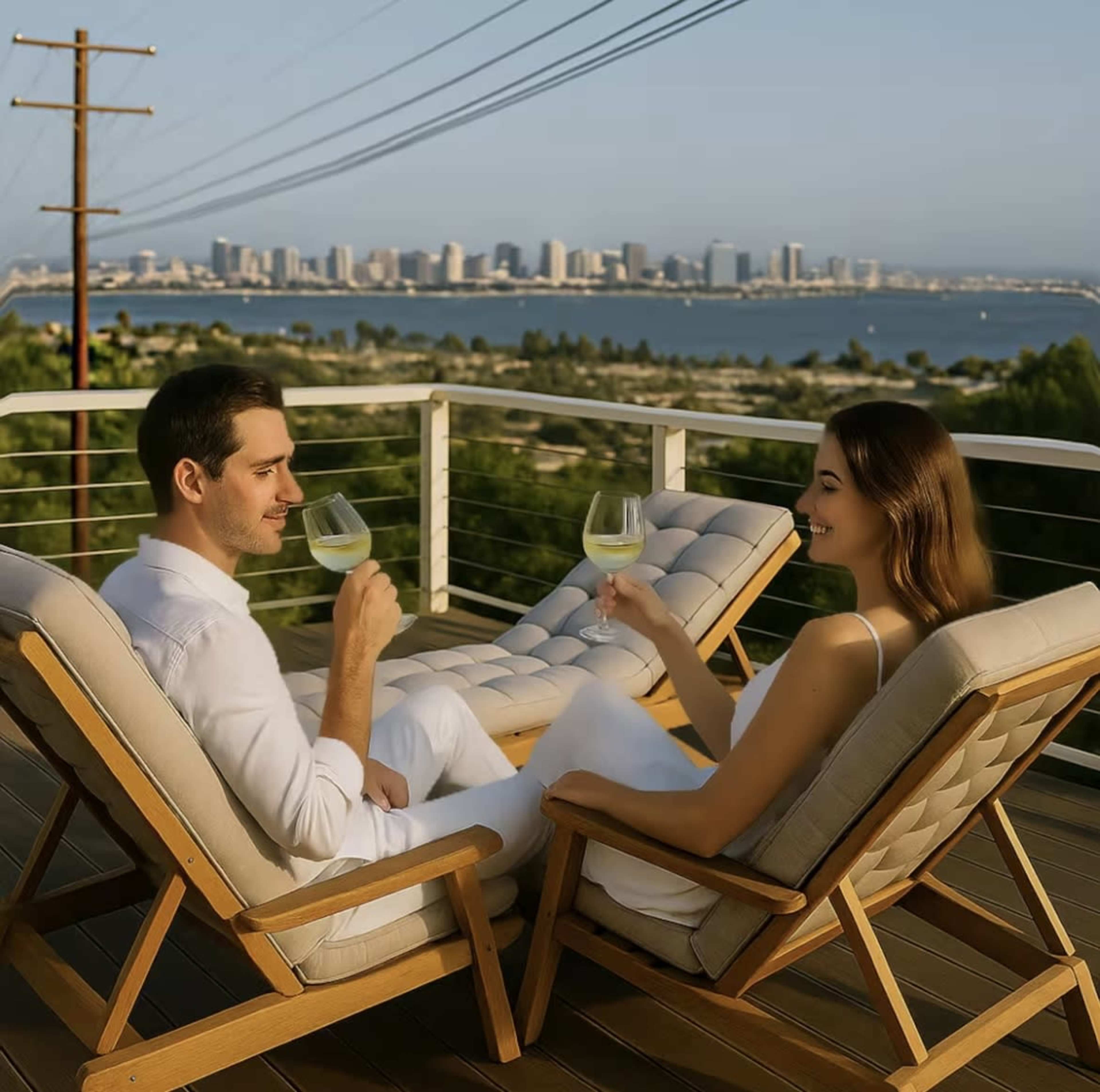 A man and a woman sit on wooden lounge chairs, holding drinks and enjoying a view of a city skyline and water in the background.