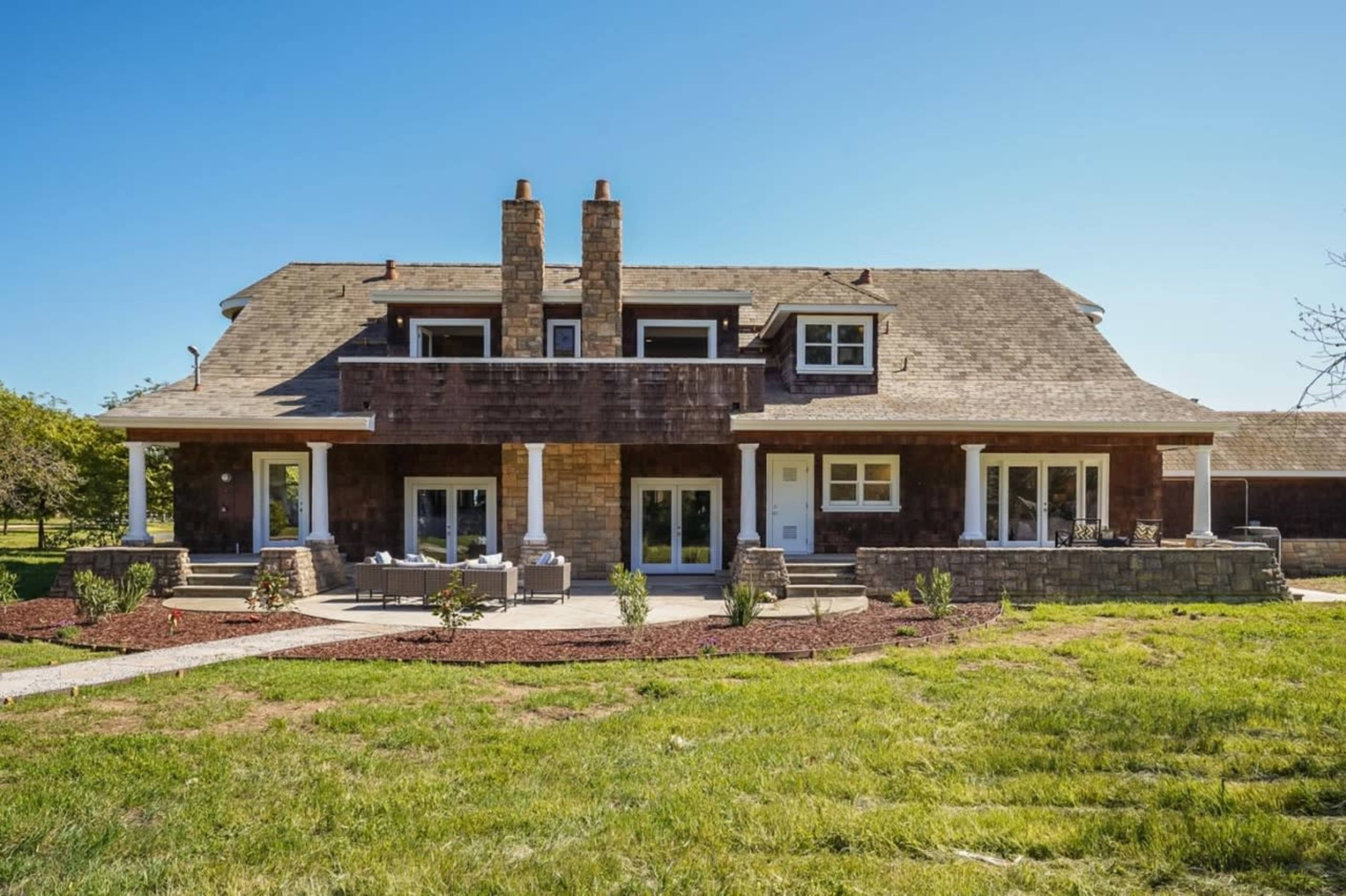 A two-story house with a brown wooden exterior and a stone patio features two chimneys and is surrounded by grass and landscaped planting beds.