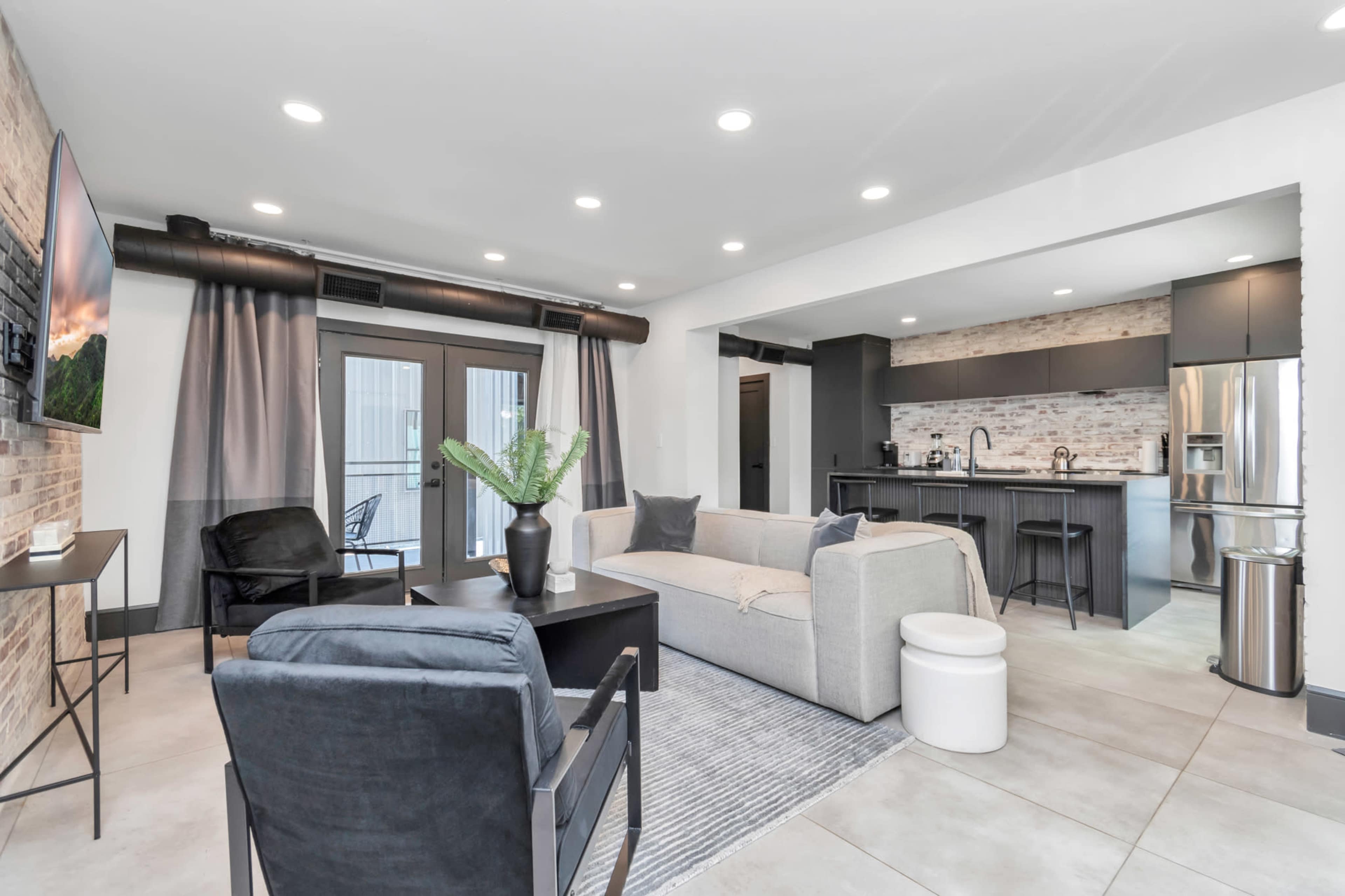 A modern living area featuring a sectional sofa, two chairs, a coffee table, and a kitchen with a brick backsplash and stainless steel appliances.