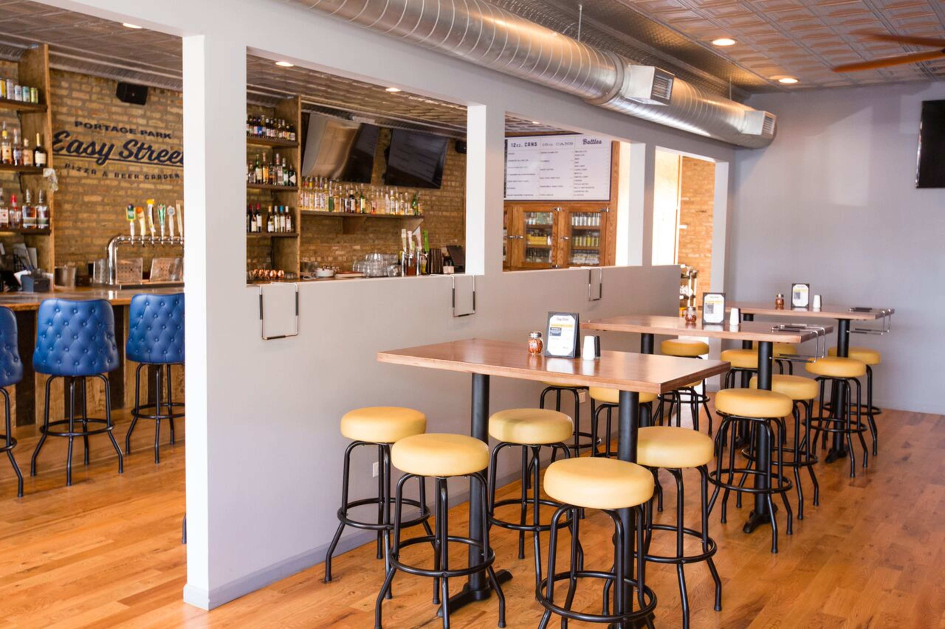 The image shows a modern bar interior featuring wooden tables, yellow stools, a brick wall, and a well-stocked bar area.