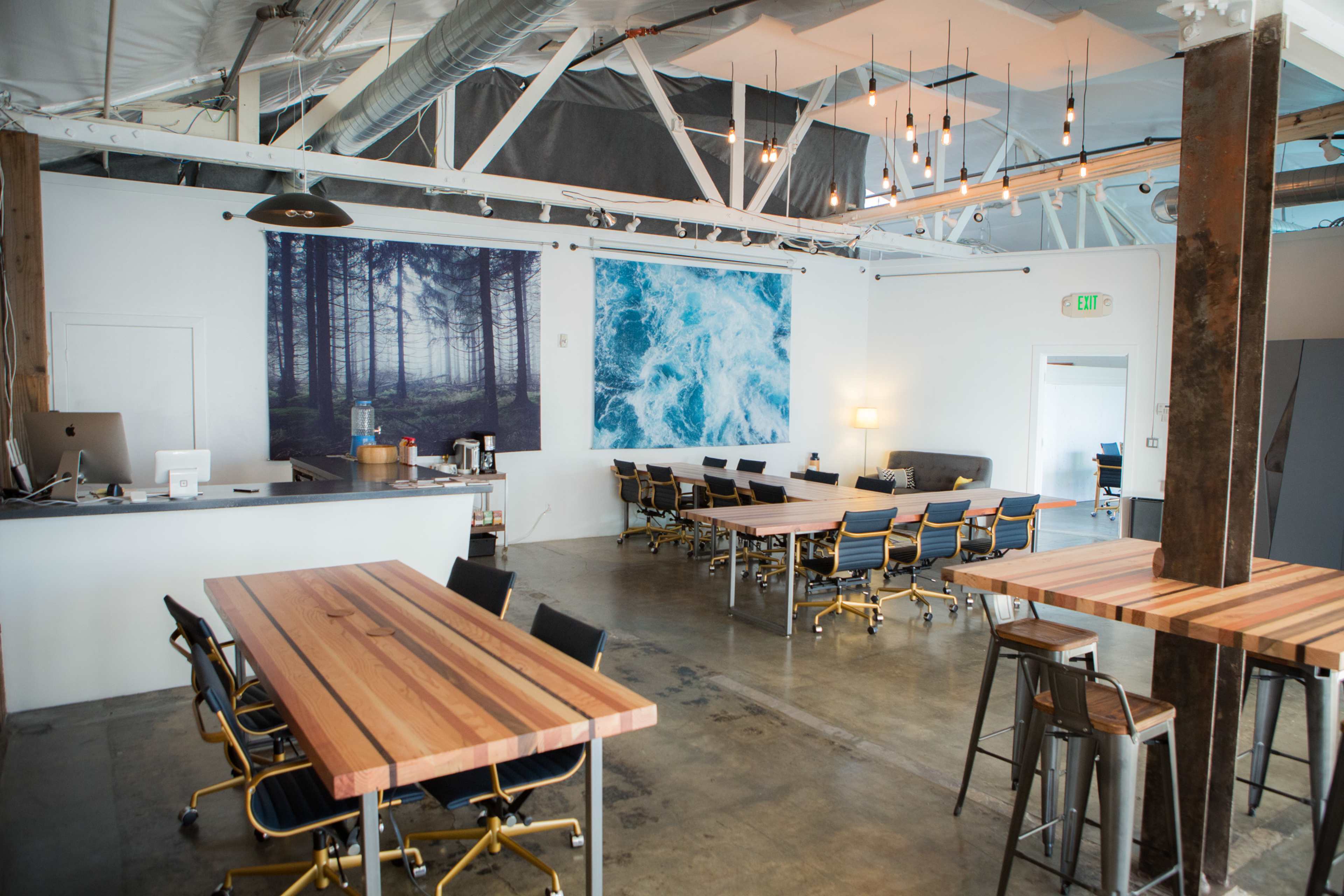 The image shows a modern workspace with wooden tables, metal chairs, and large wall art depicting a forest and ocean.