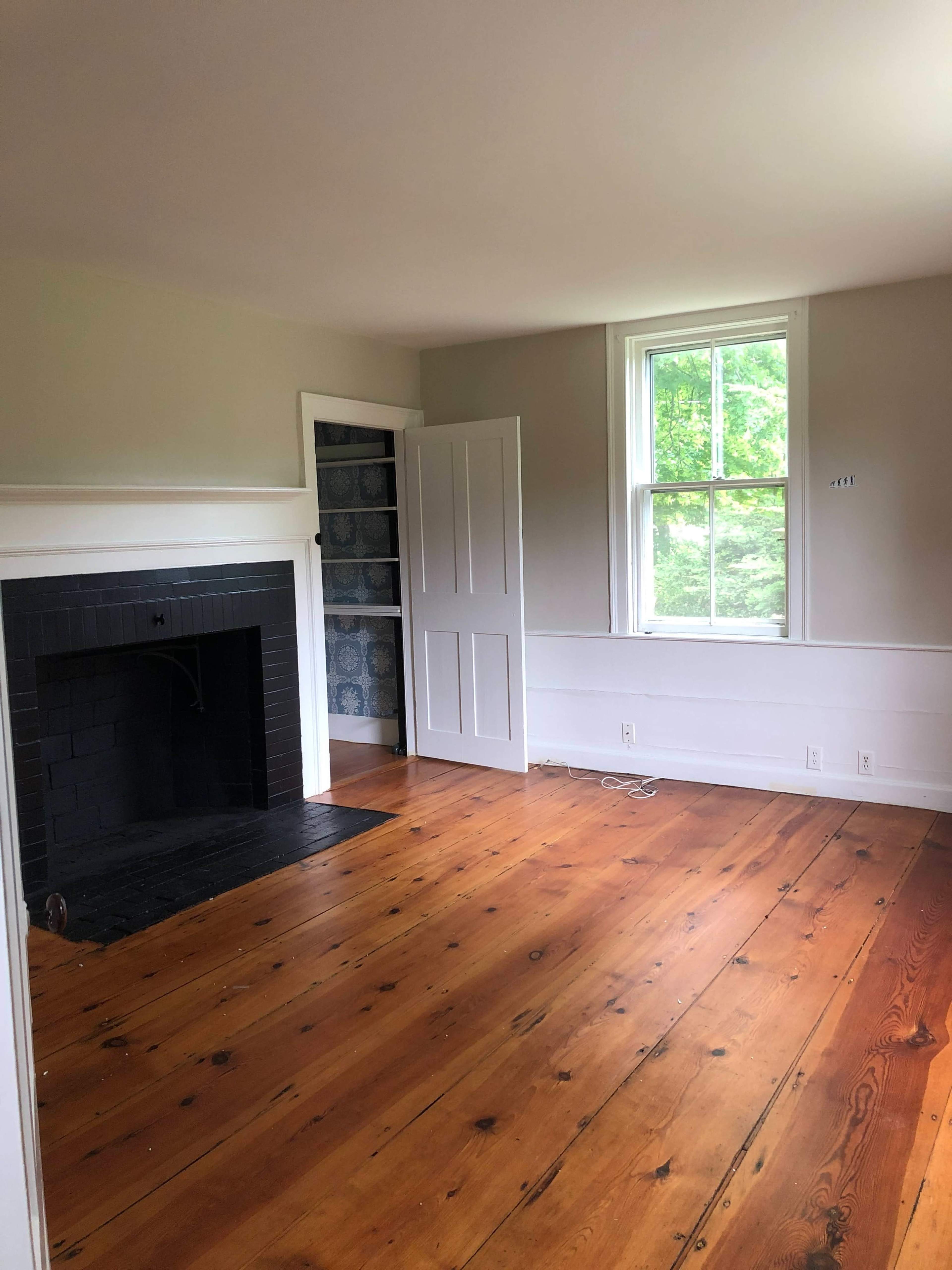 An empty room with wooden floors, a fireplace, a window, and a partially open door.