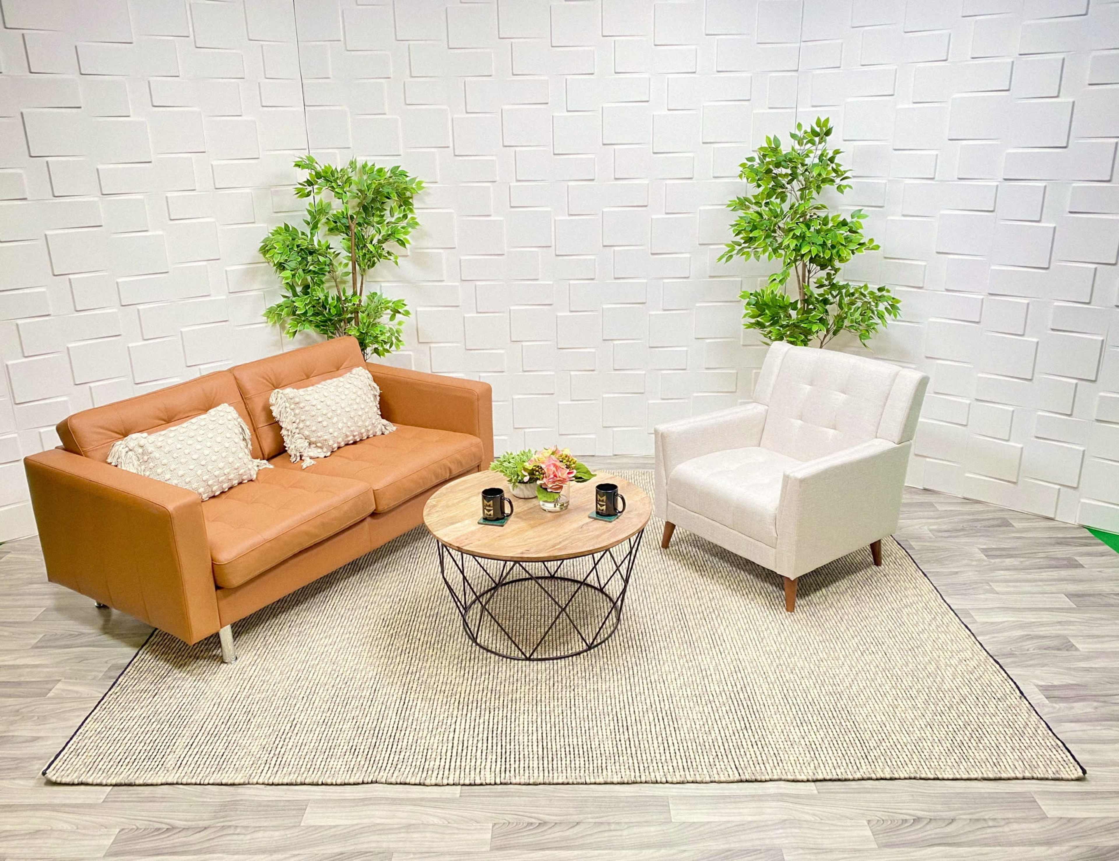 A modern living space featuring a tan sofa, a light-colored armchair, a round wooden coffee table, and artificial plants against a textured white wall.