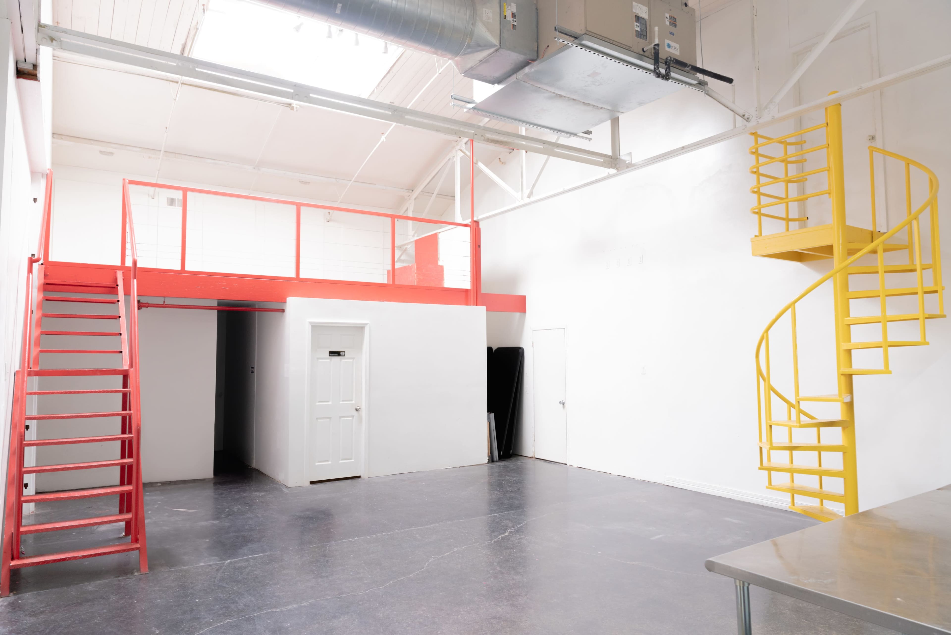 The image shows an industrial-style interior space featuring a red staircase on the left leading to a loft area, a yellow spiral staircase on the right, and white walls with polished concrete flooring.