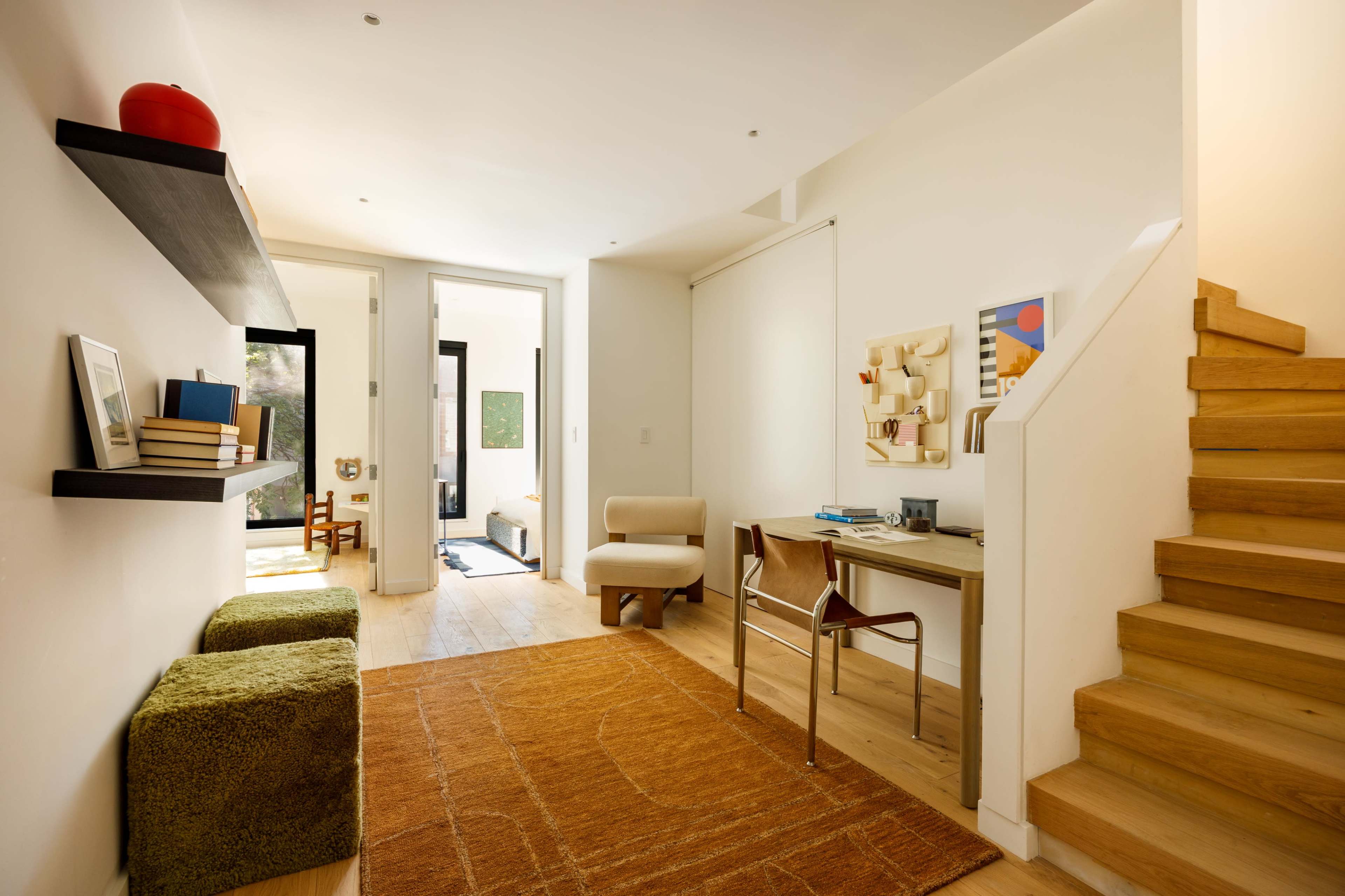 The image shows a modern entryway featuring wooden stairs, a small desk with a chair, and a cozy seating area with green ottomans on a large rug.