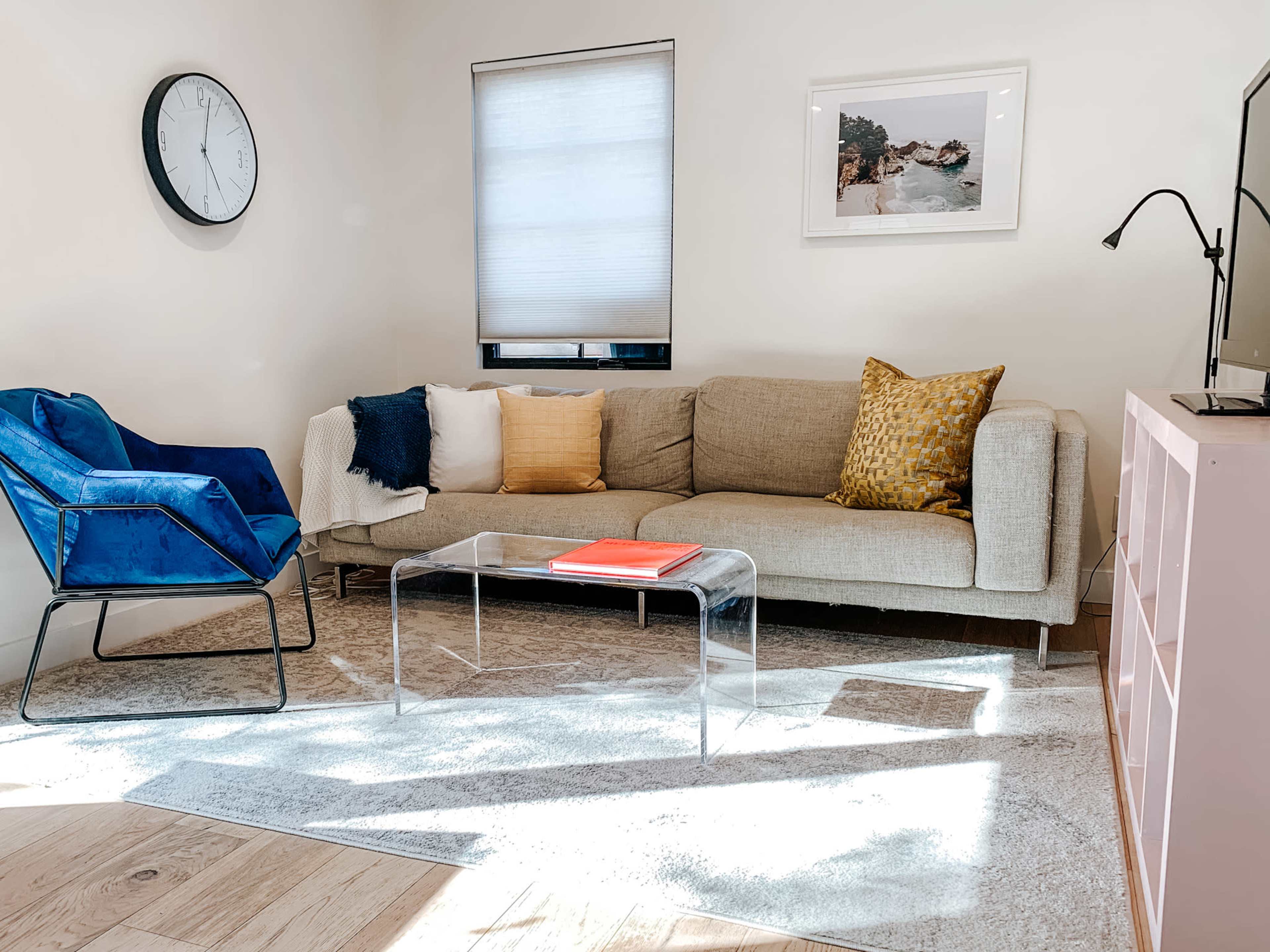 The image shows a modern living room with a light-colored sofa, a clear coffee table, a blue accent chair, and a wall clock.