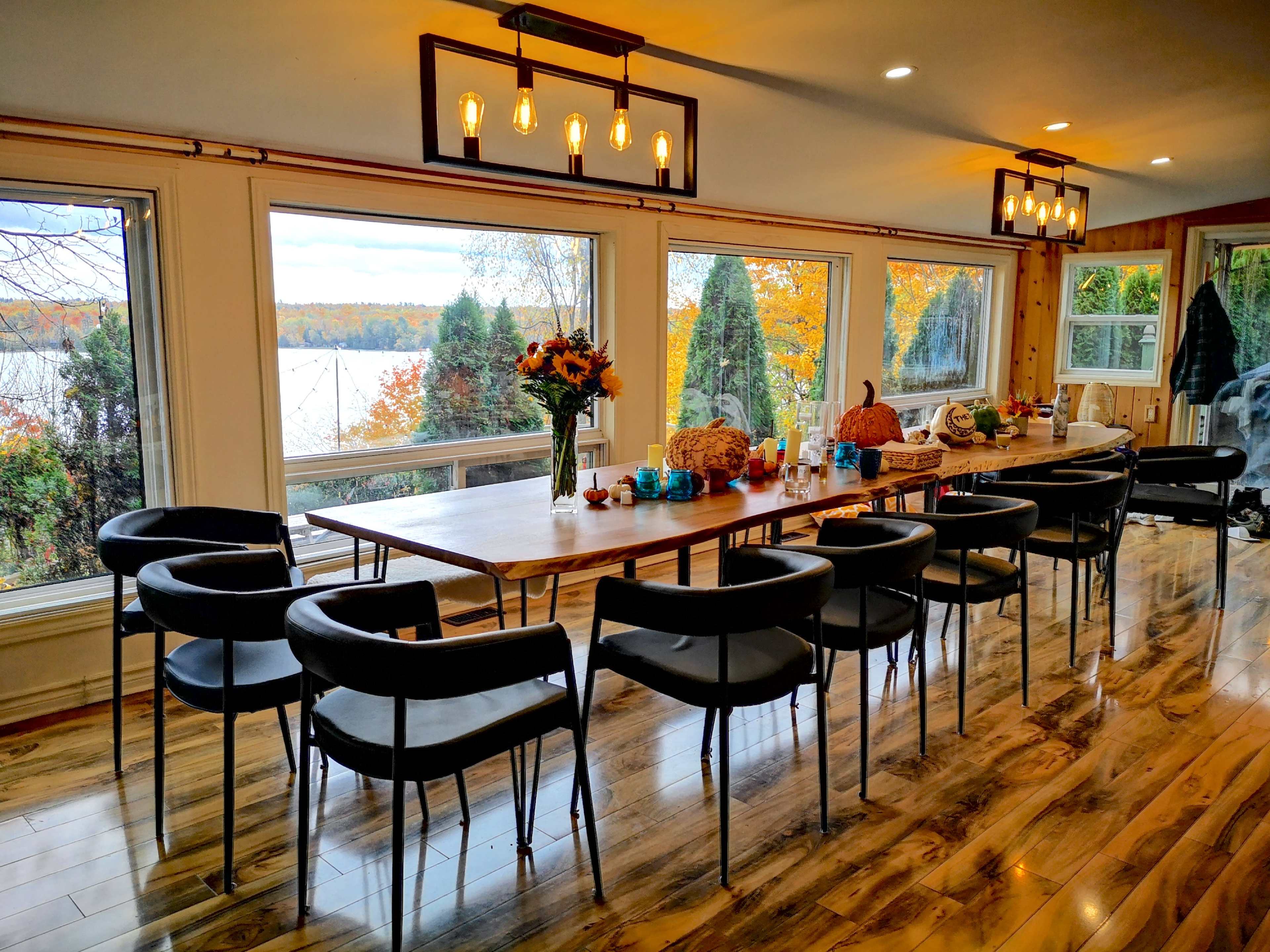 A long table with black chairs is set up near large windows overlooking a colorful autumn landscape by a lake.