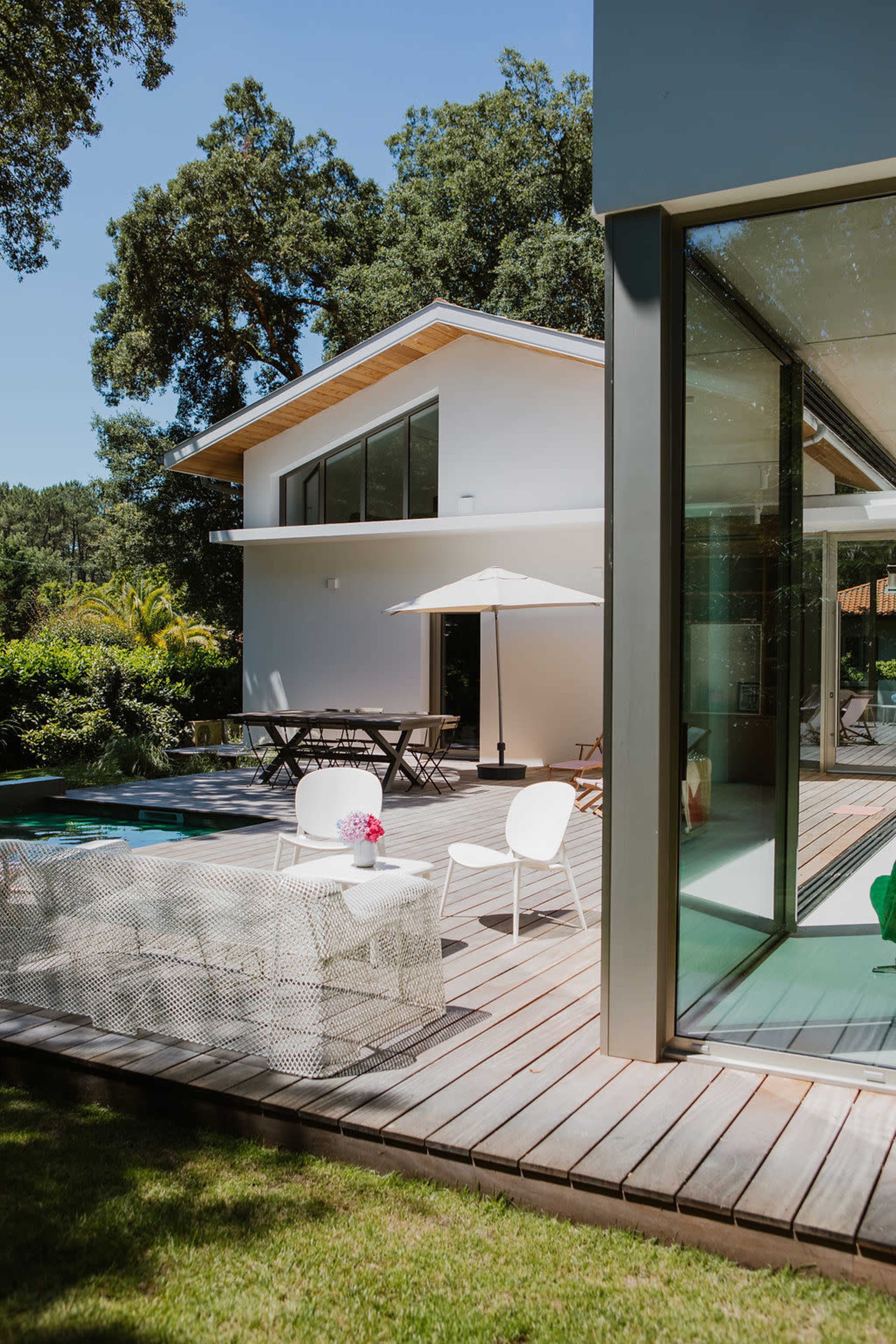 The image shows a modern house with a wooden deck, outdoor seating, and a swimming pool surrounded by greenery.