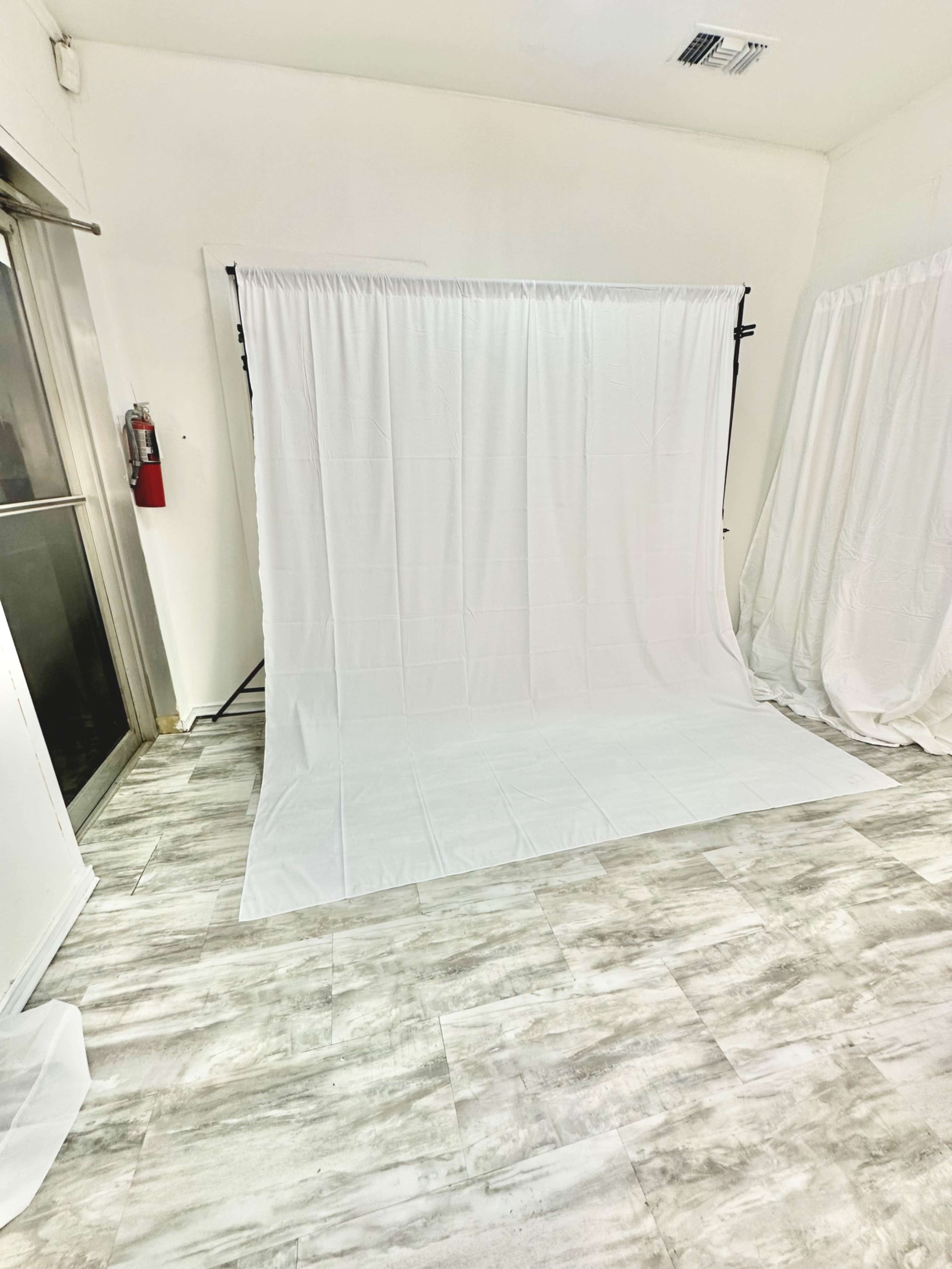 A room with a white backdrop set up on a stand, surrounded by white curtains and featuring a light-colored tiled floor.