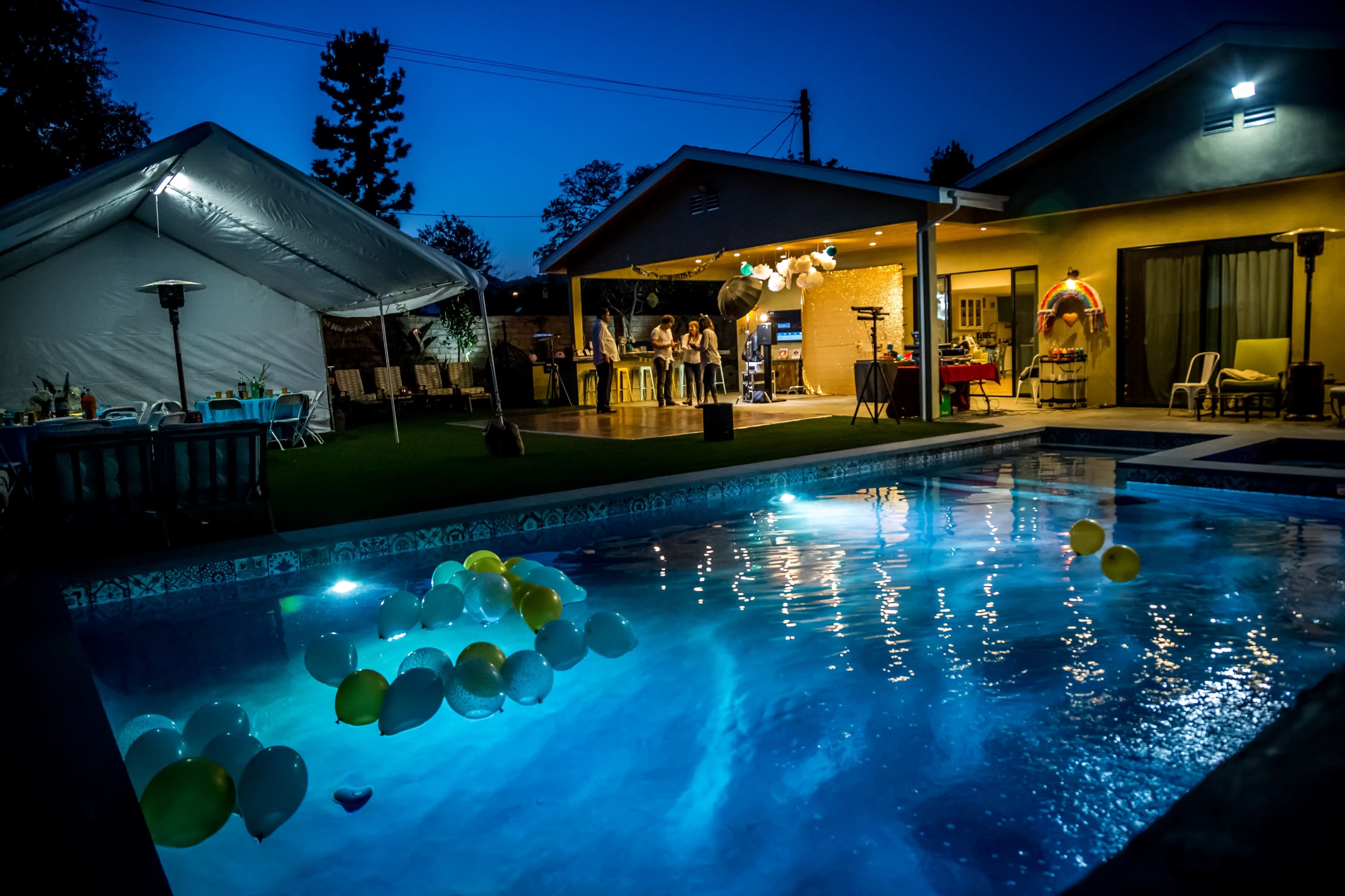 A backyard party scene features a pool illuminated by balloons and a large tent set up next to a house at twilight.