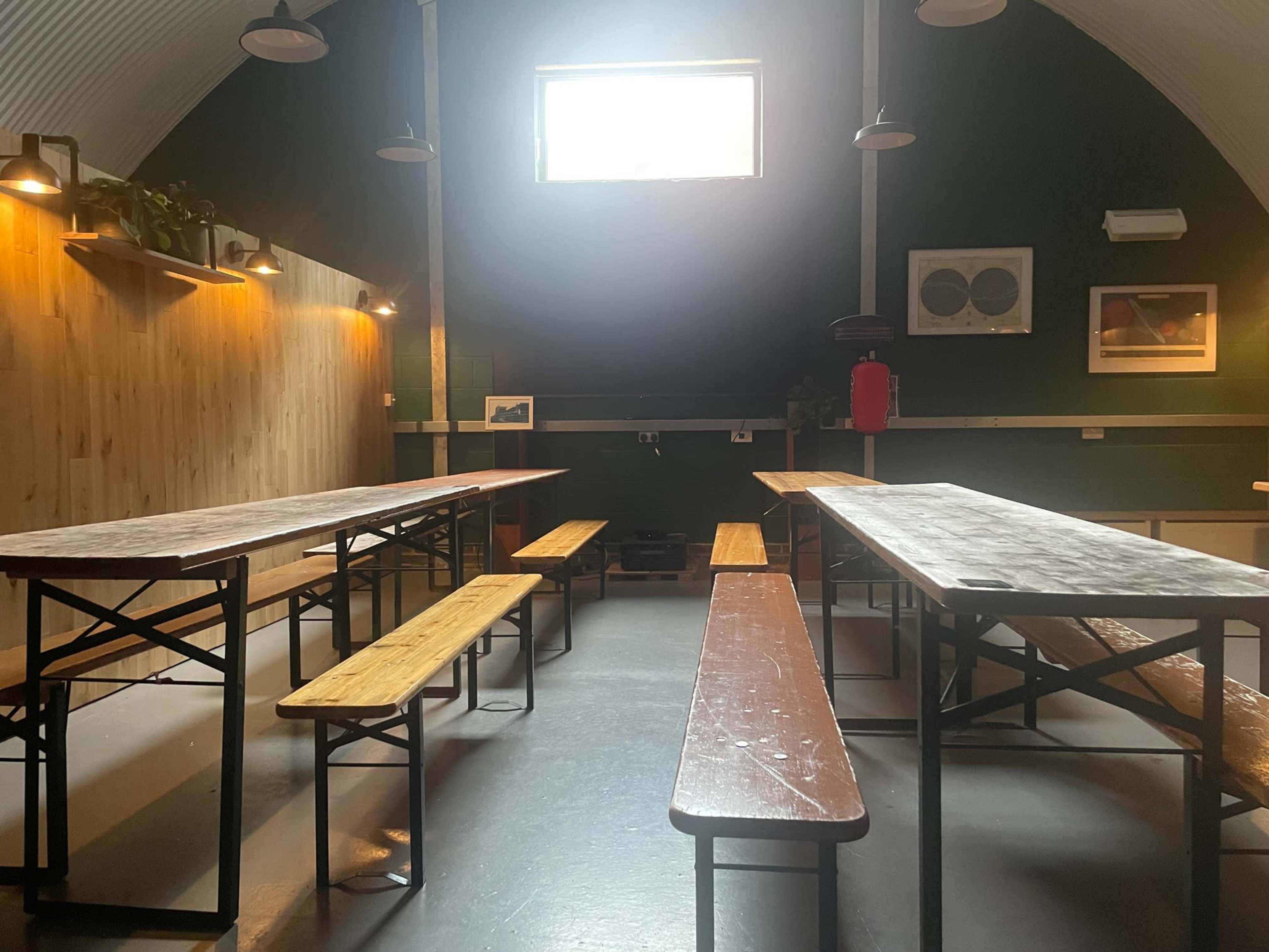 The image shows a spacious room with wooden benches and tables arranged, illuminated by natural light coming through a window.