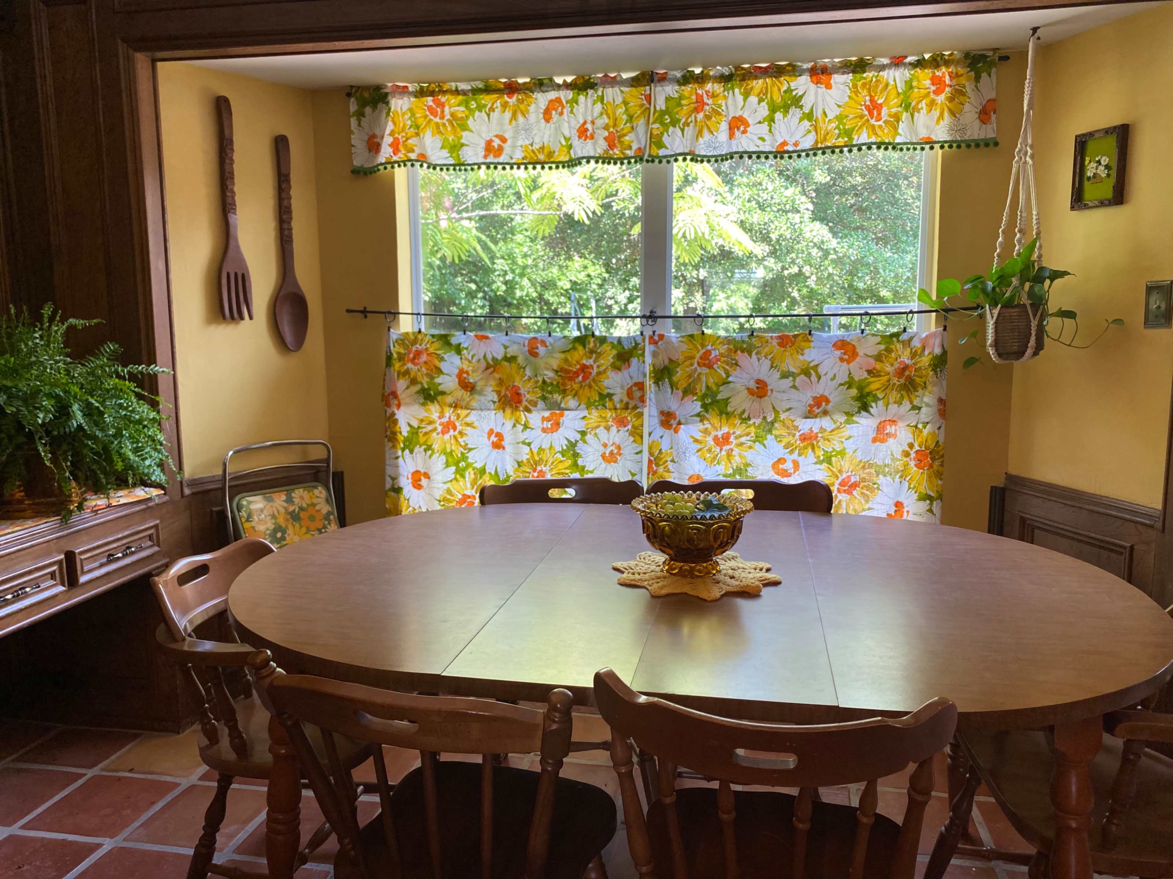 A dining room features a round wooden table surrounded by chairs, with floral-patterned curtains covering a window that overlooks greenery.