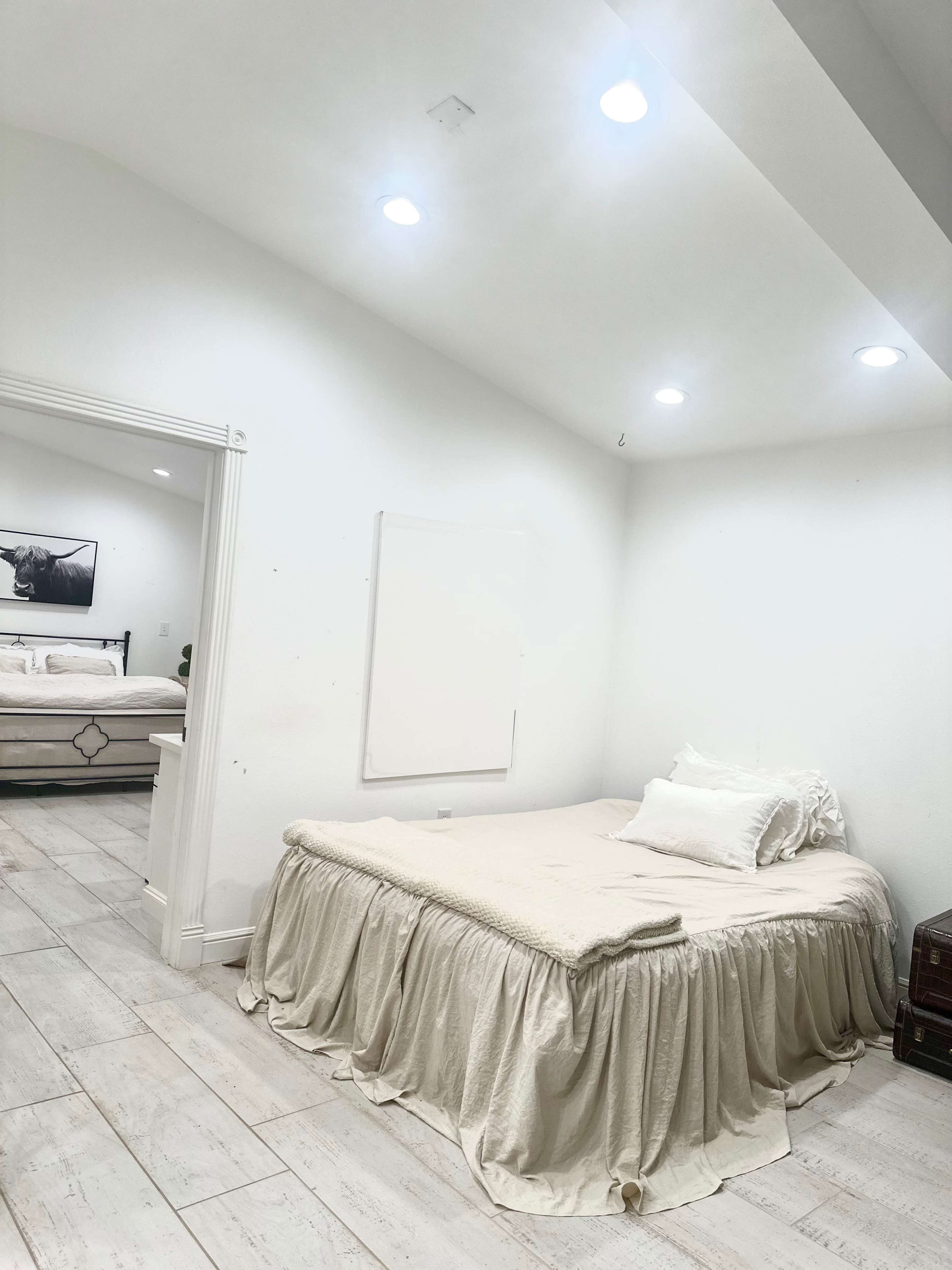 A minimally furnished bedroom with a light-colored bedspread and white walls, featuring recessed lighting and a doorway leading to another room.