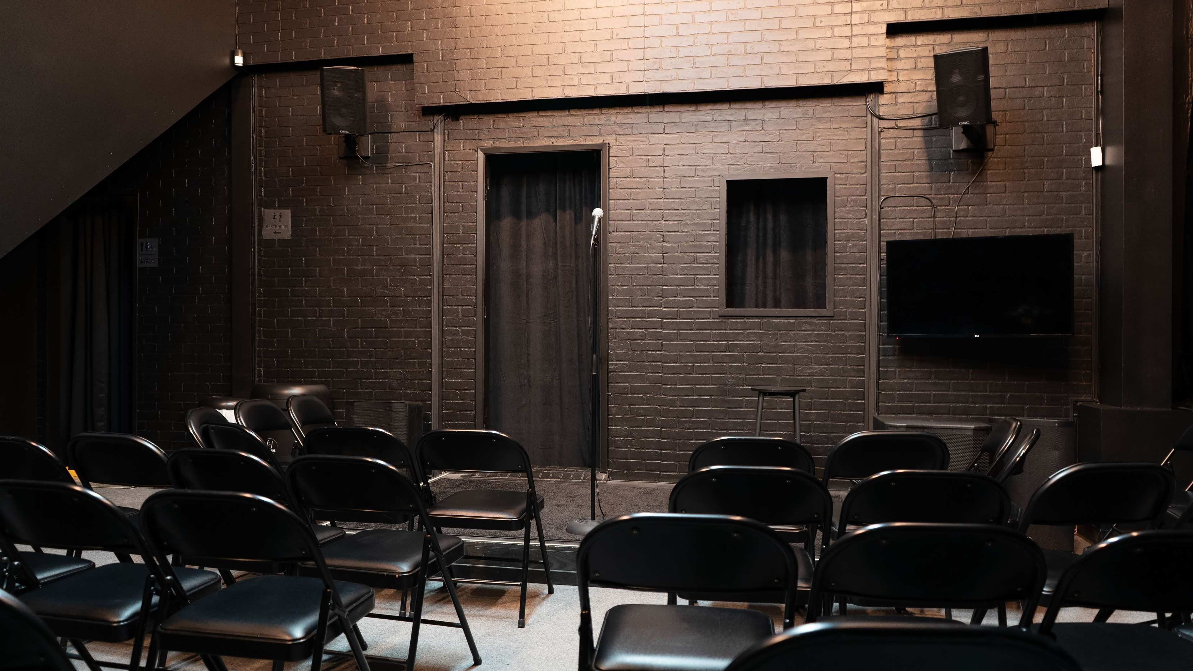 A dimly lit comedy stage features a microphone stand and a backdrop of black brick, with rows of black folding chairs arranged in front.