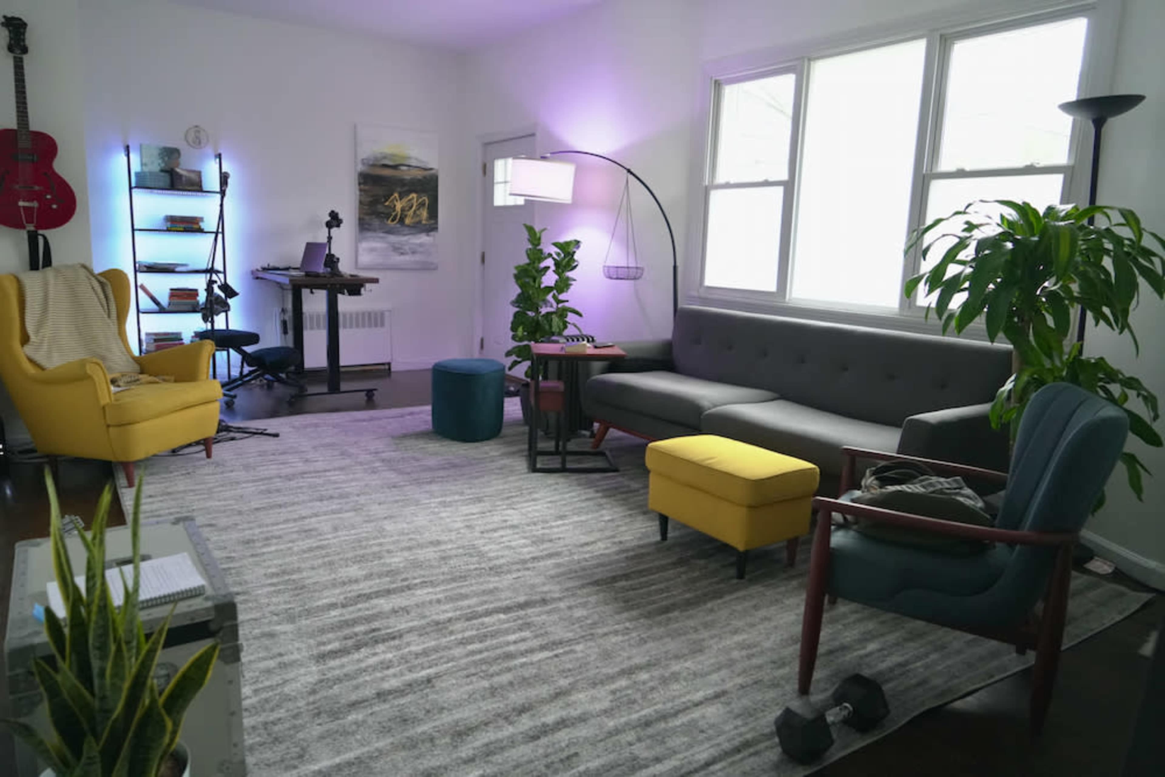 A modern living room with a gray sectional sofa, yellow accent chair, green plants, and a desk setup in the corner.