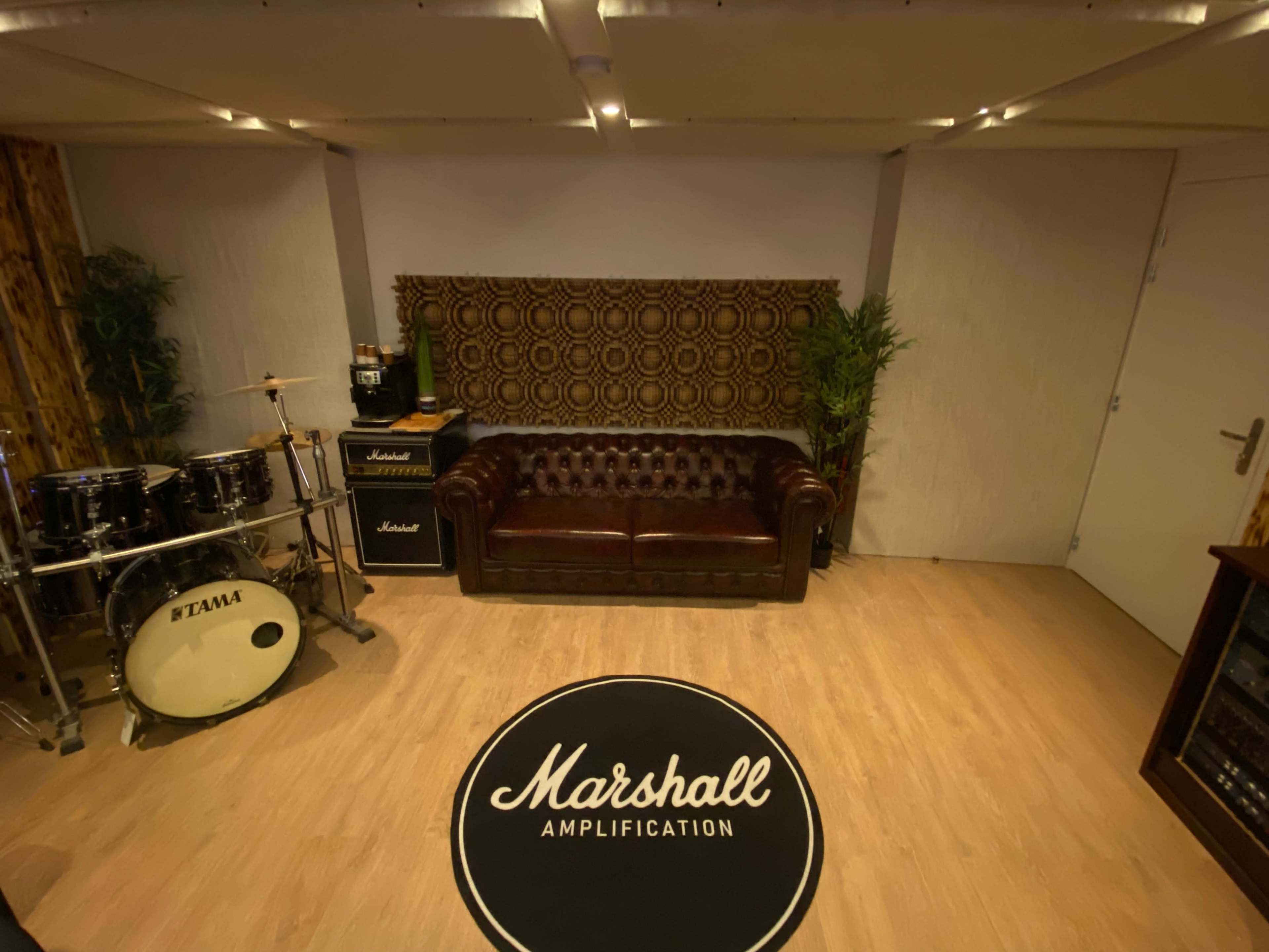 A music rehearsal space featuring a drum set, an amplifier stack, a brown leather couch, and a circular rug displaying the "Marshall Amplification" logo.
