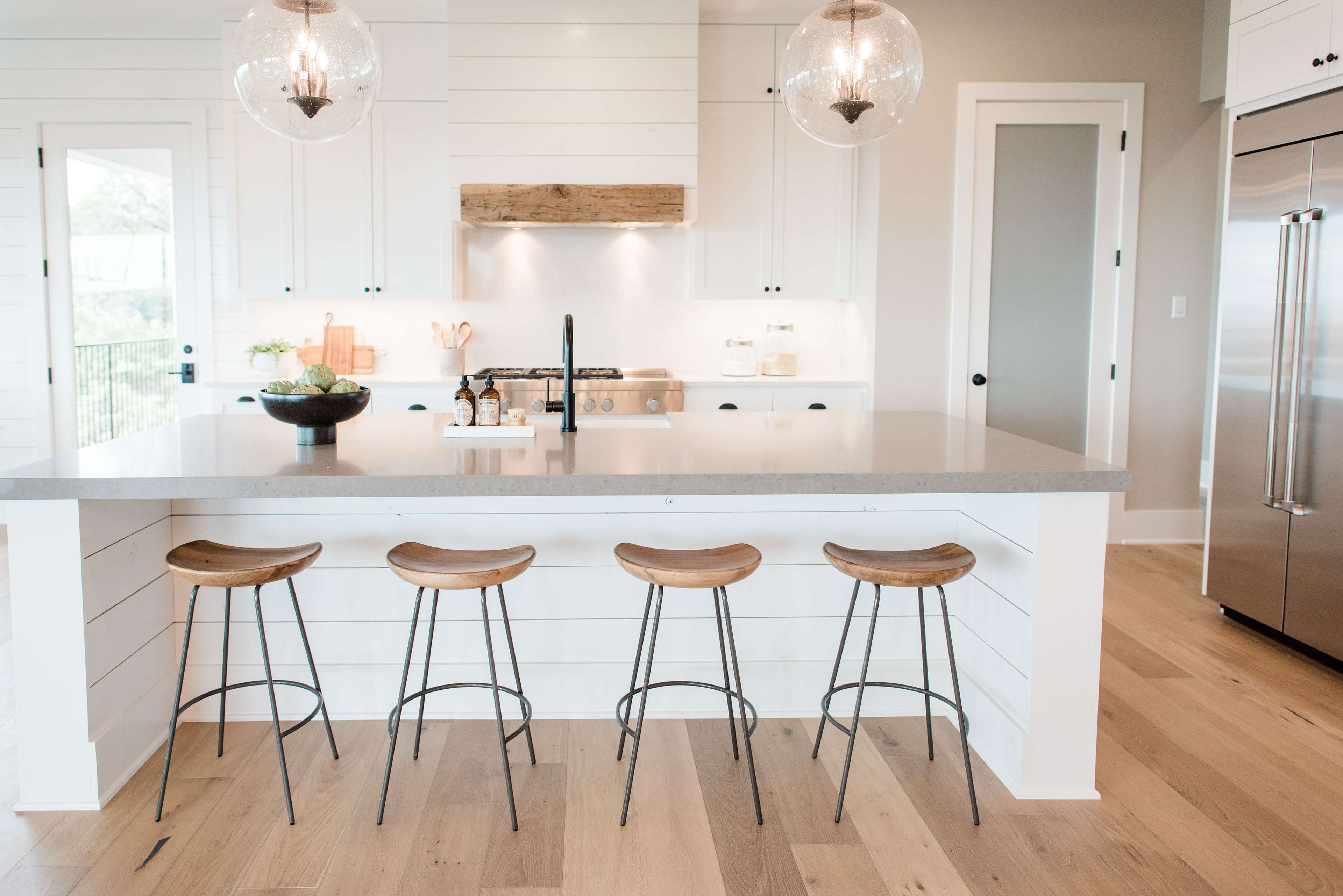 A modern kitchen featuring a long countertop, four wooden stools, and pendant lighting over a cooking area.