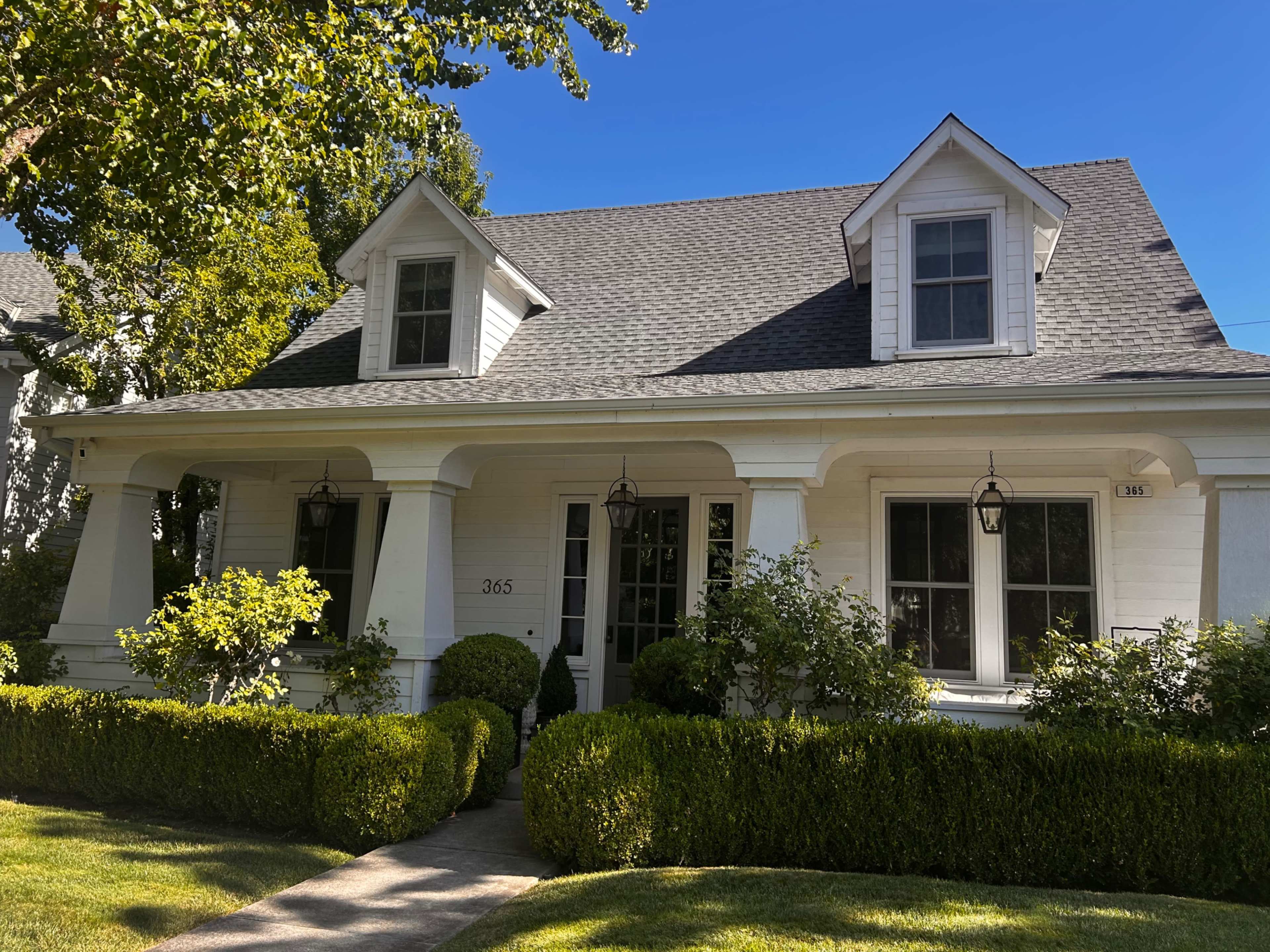 A white house with a sloped roof and two front gables is framed by neatly trimmed hedges and a stone walkway leading to the entrance.