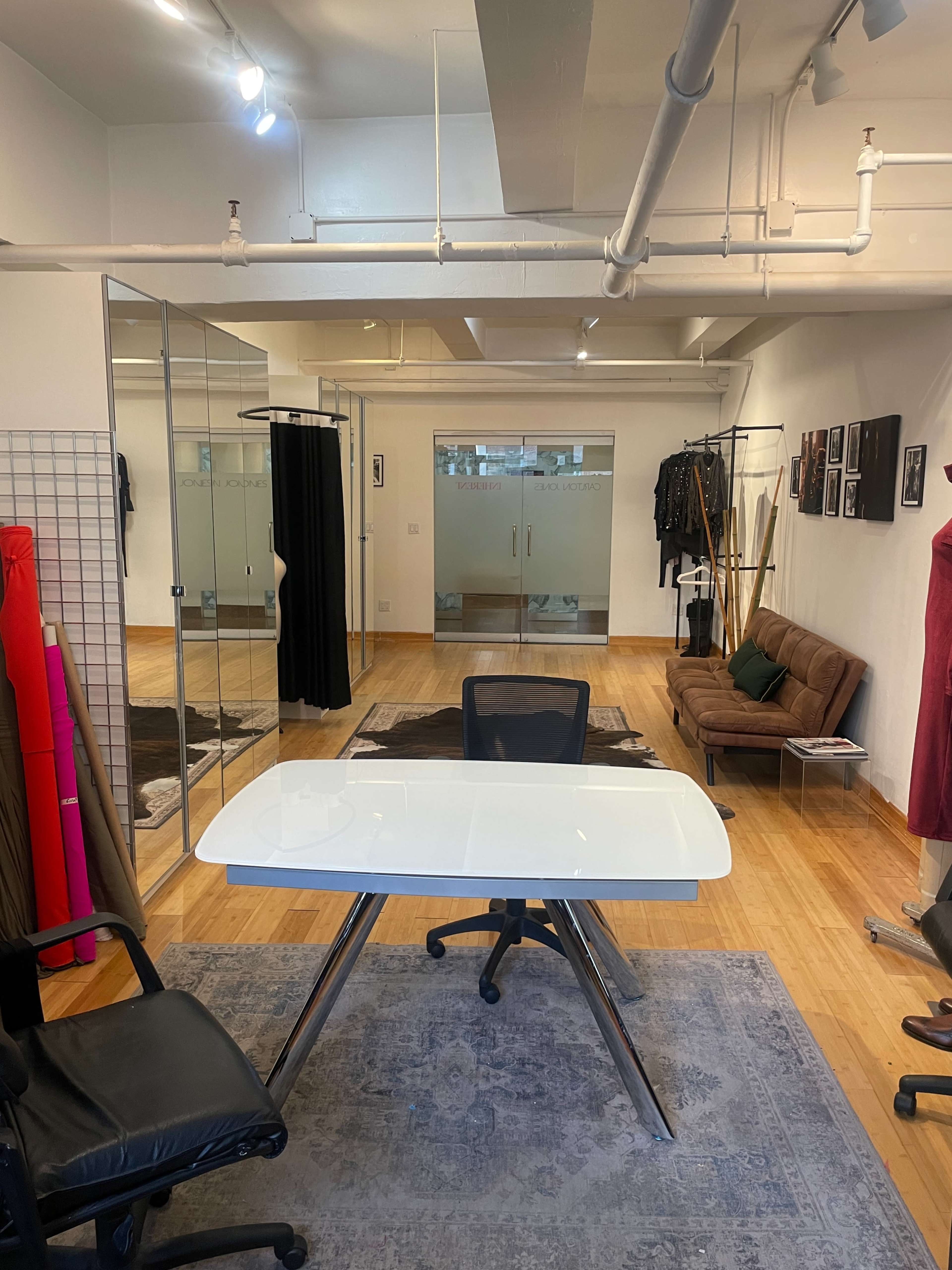 The image shows a modern office space featuring a white desk in the foreground, with a brown couch and clothing racks in the background.
