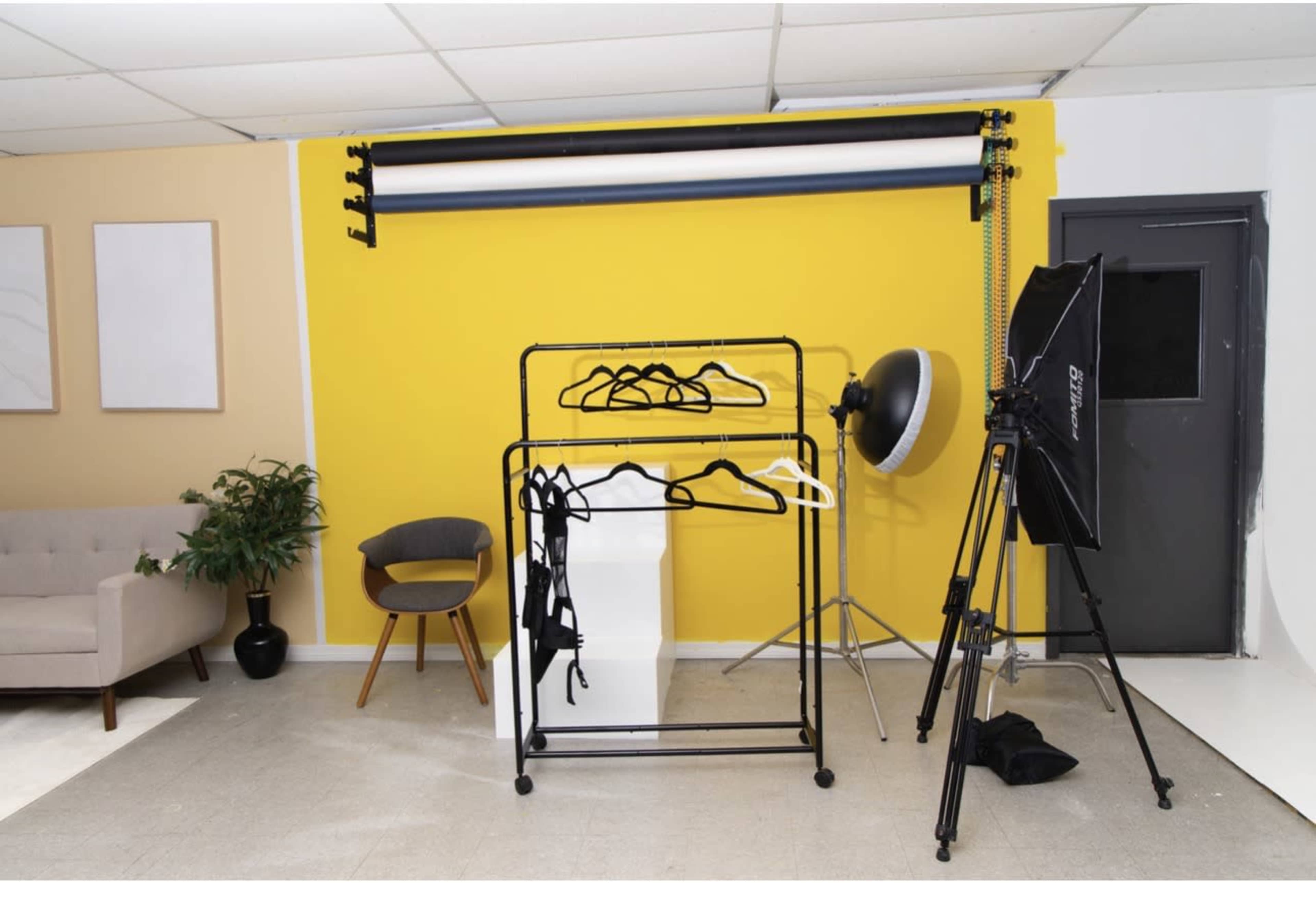 The image shows a studio space with a yellow wall, a clothing rack with garments, a softbox light, a gray door, and a couch with a plant nearby.