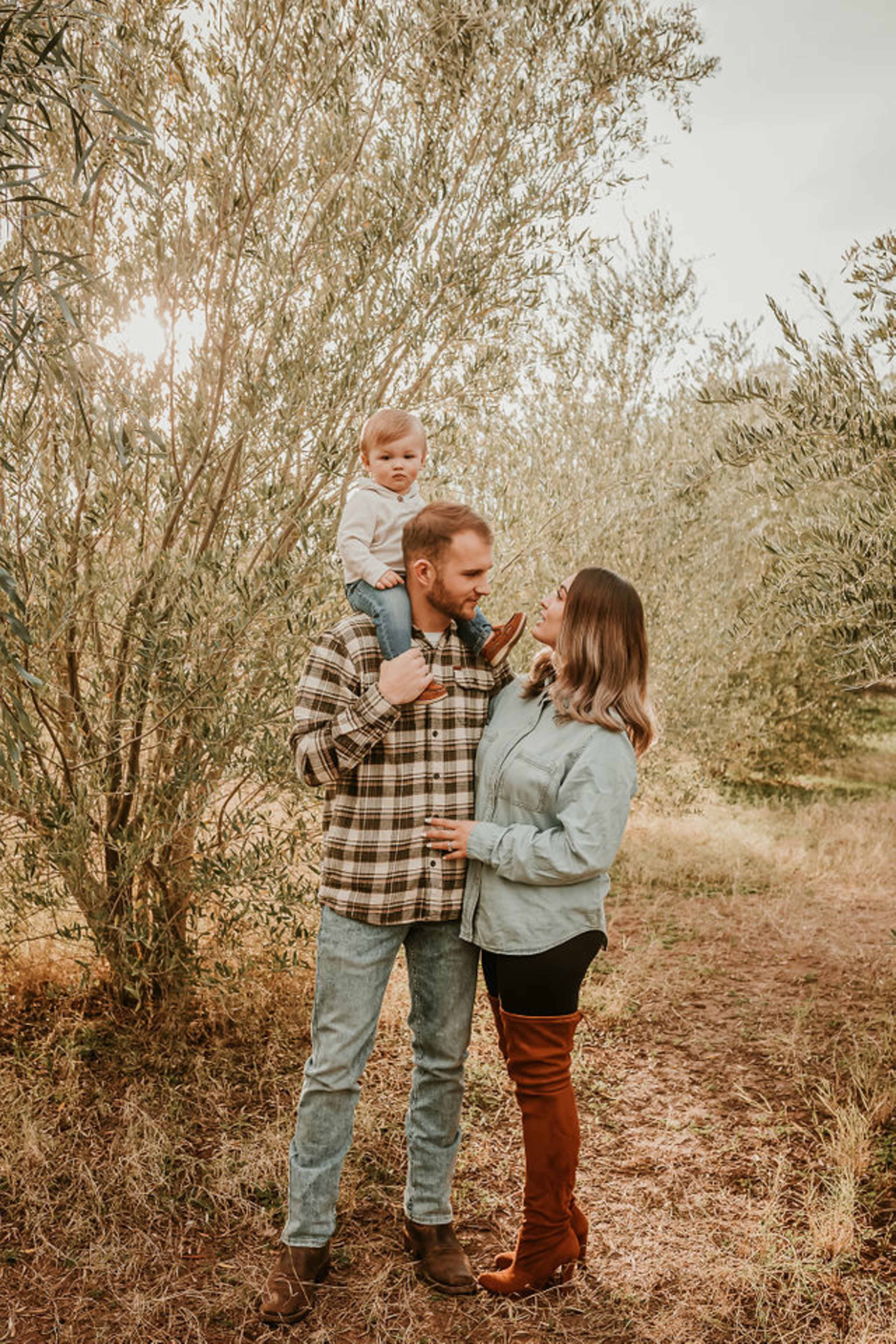 A family of three is standing in an olive grove, with a father holding a toddler on his shoulders while the mother leans in close.