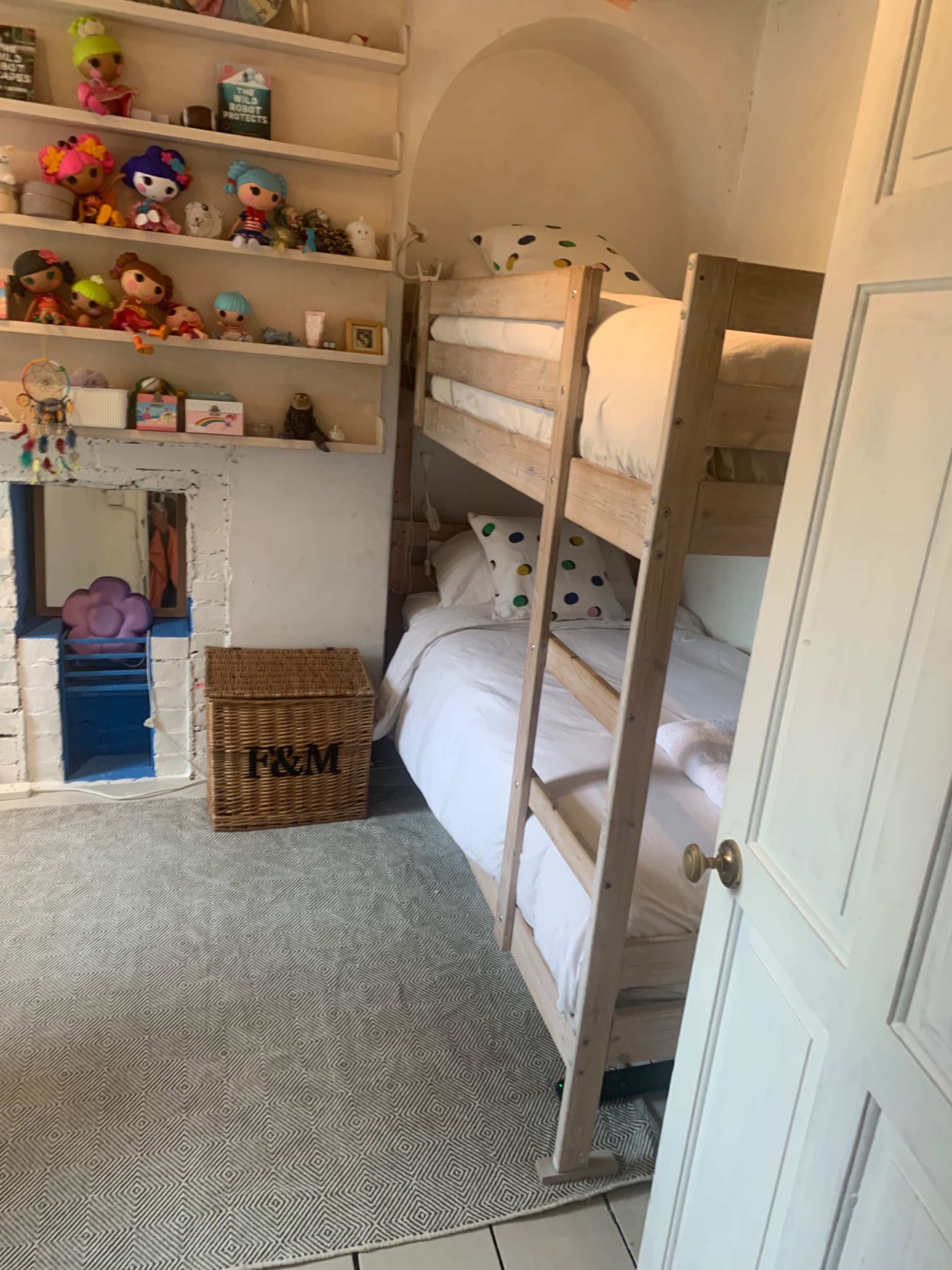 The image shows a cozy children's bedroom with a bunk bed, a doll shelf above, and a woven storage basket near a blue-painted fireplace.