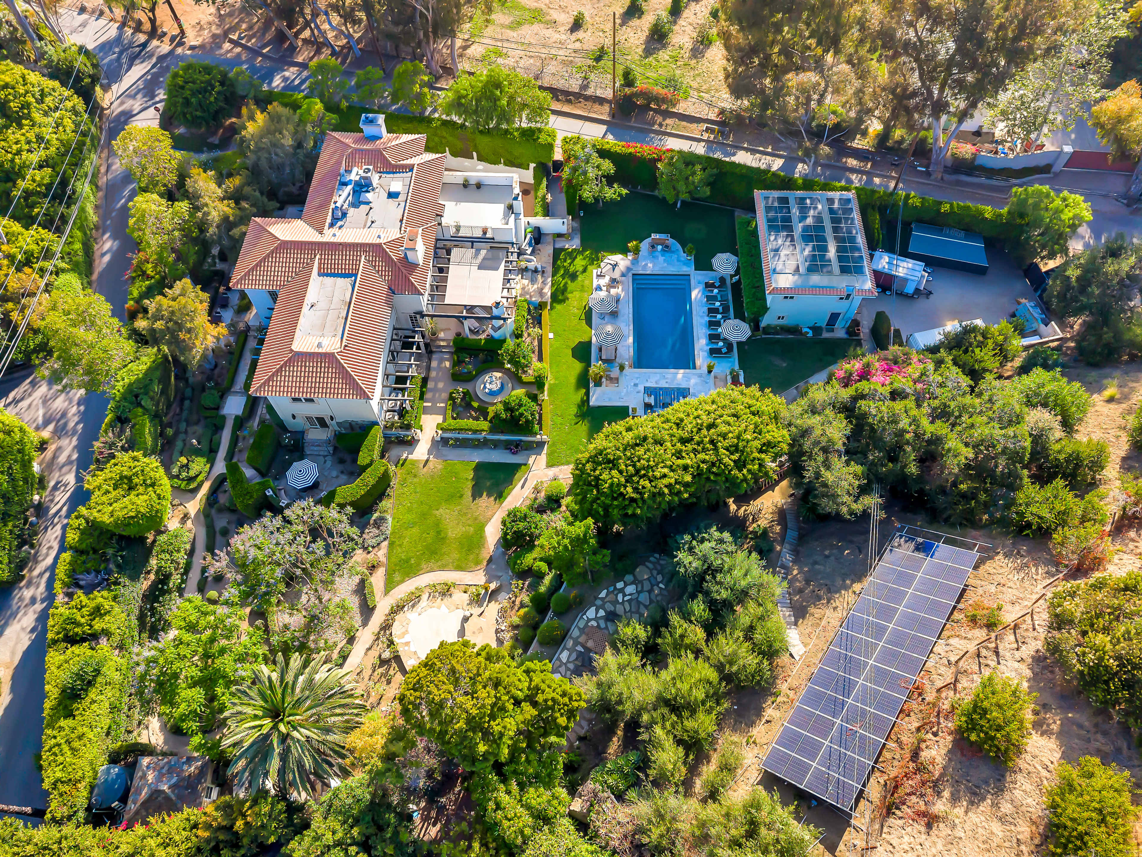 The image shows an aerial view of a residential area featuring two large houses, a swimming pool, landscaped gardens, and solar panels on the ground.