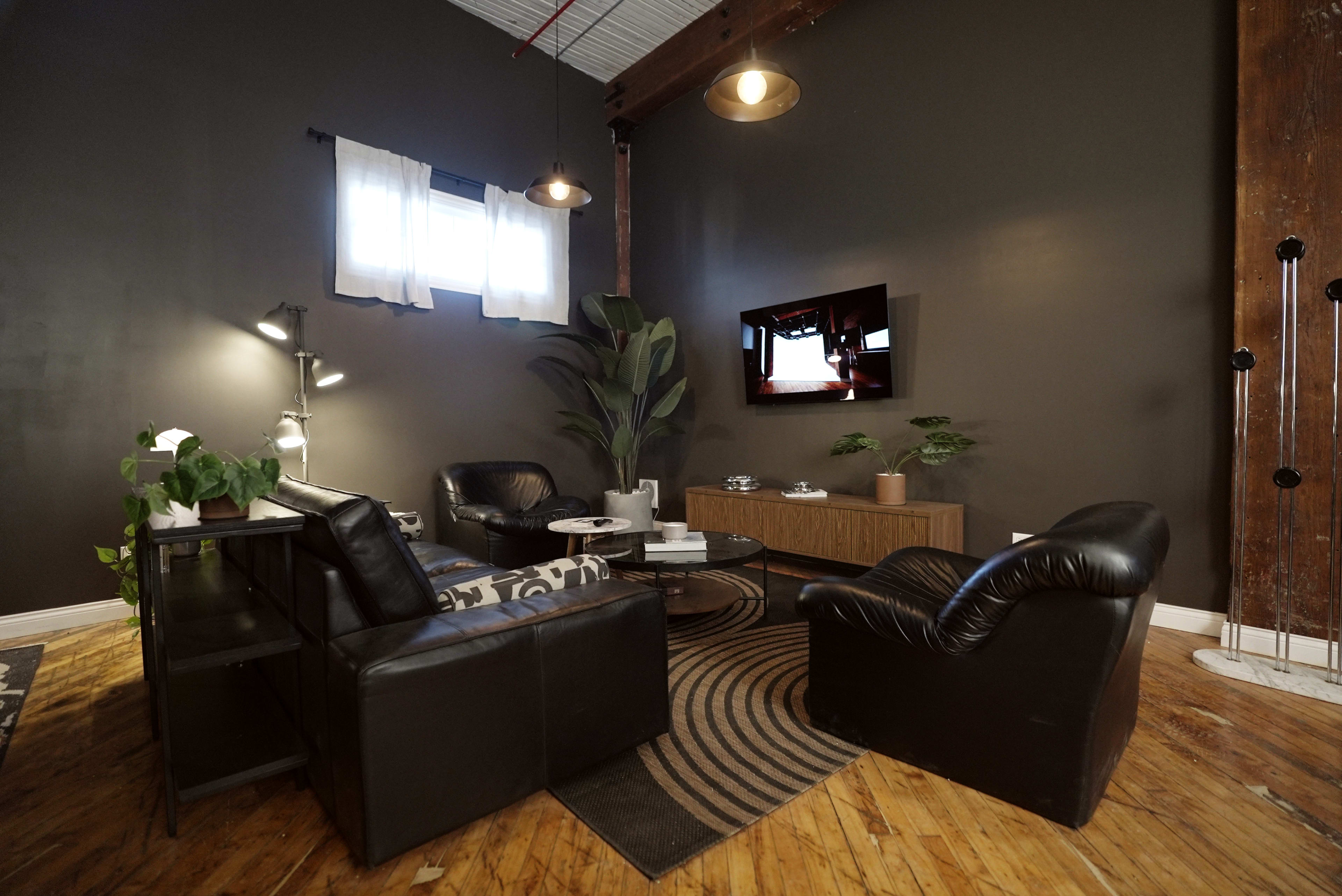 The image shows a modern living room with dark walls, wooden flooring, two leather chairs, a couch, and a television mounted on the wall.
